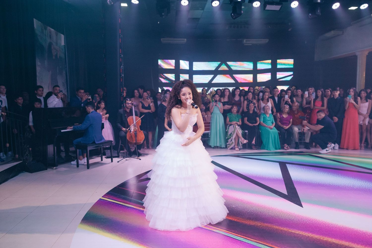 A bride in a white ruffled ball gown stands on a colorfully lit dance floor at an elegant wedding reception.
