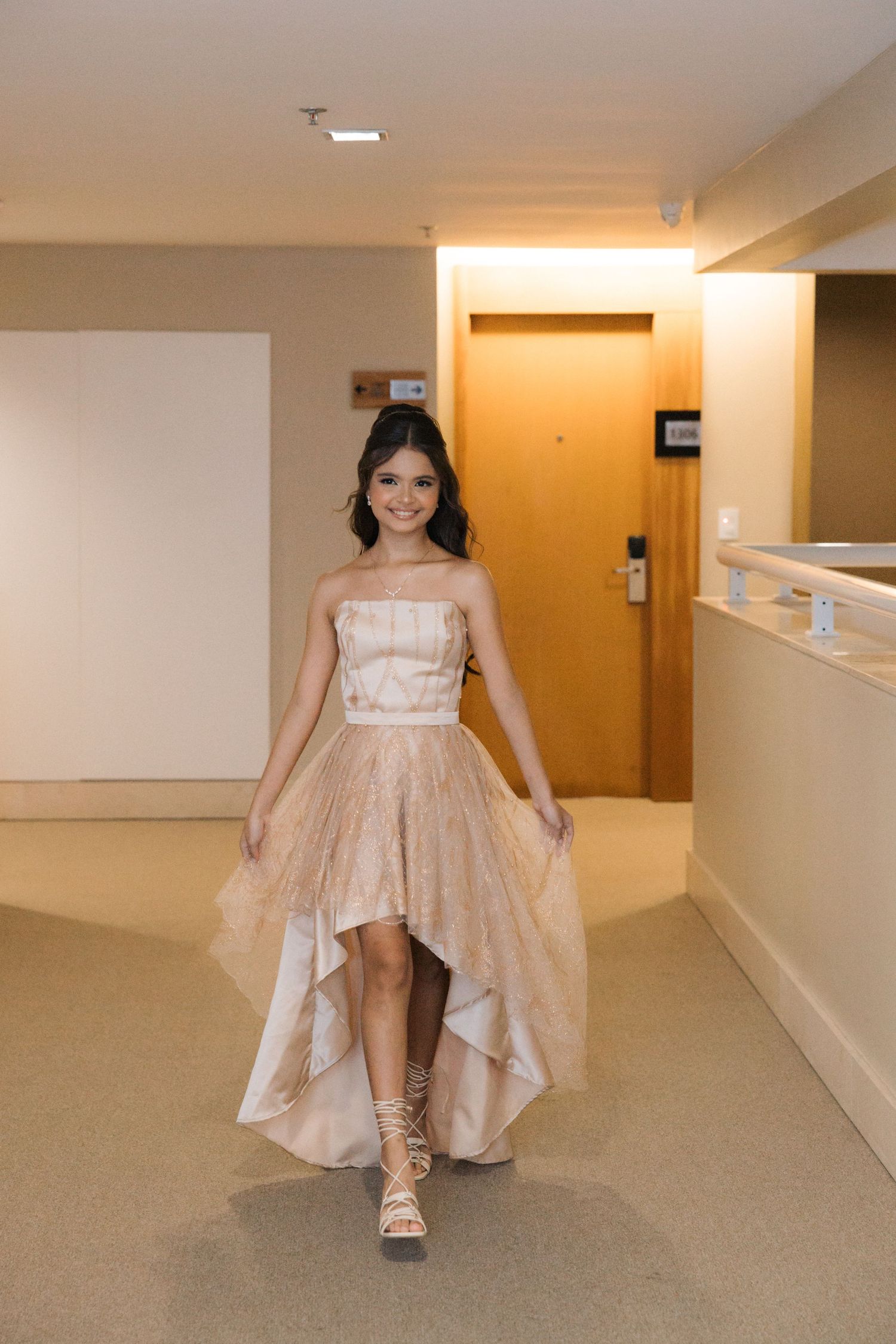 A person in an elegant champagne-colored high-low gown poses in a hotel hallway.
