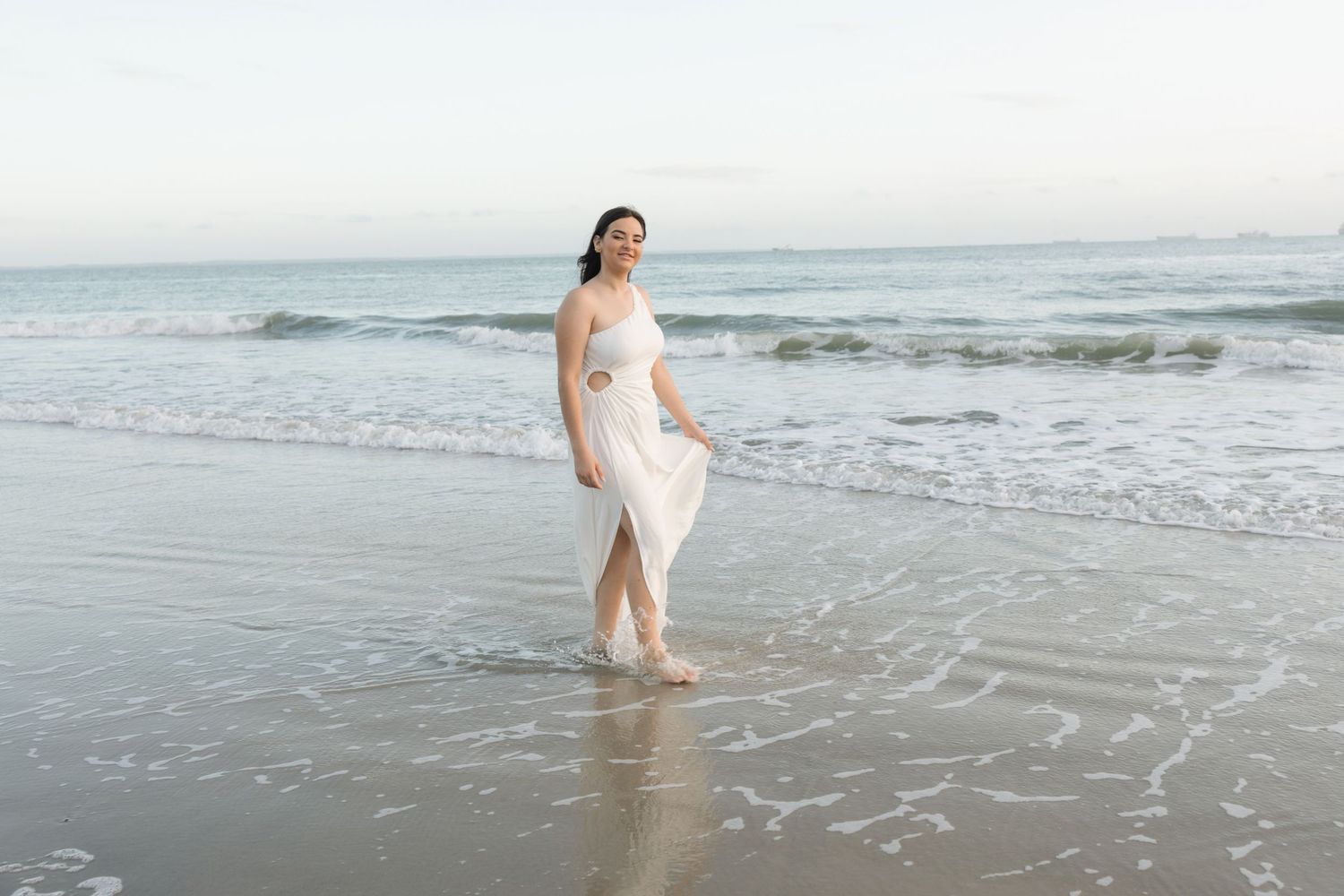 A woman in a flowing white dress walks along the shoreline at sunset as gentle waves roll onto the beach.