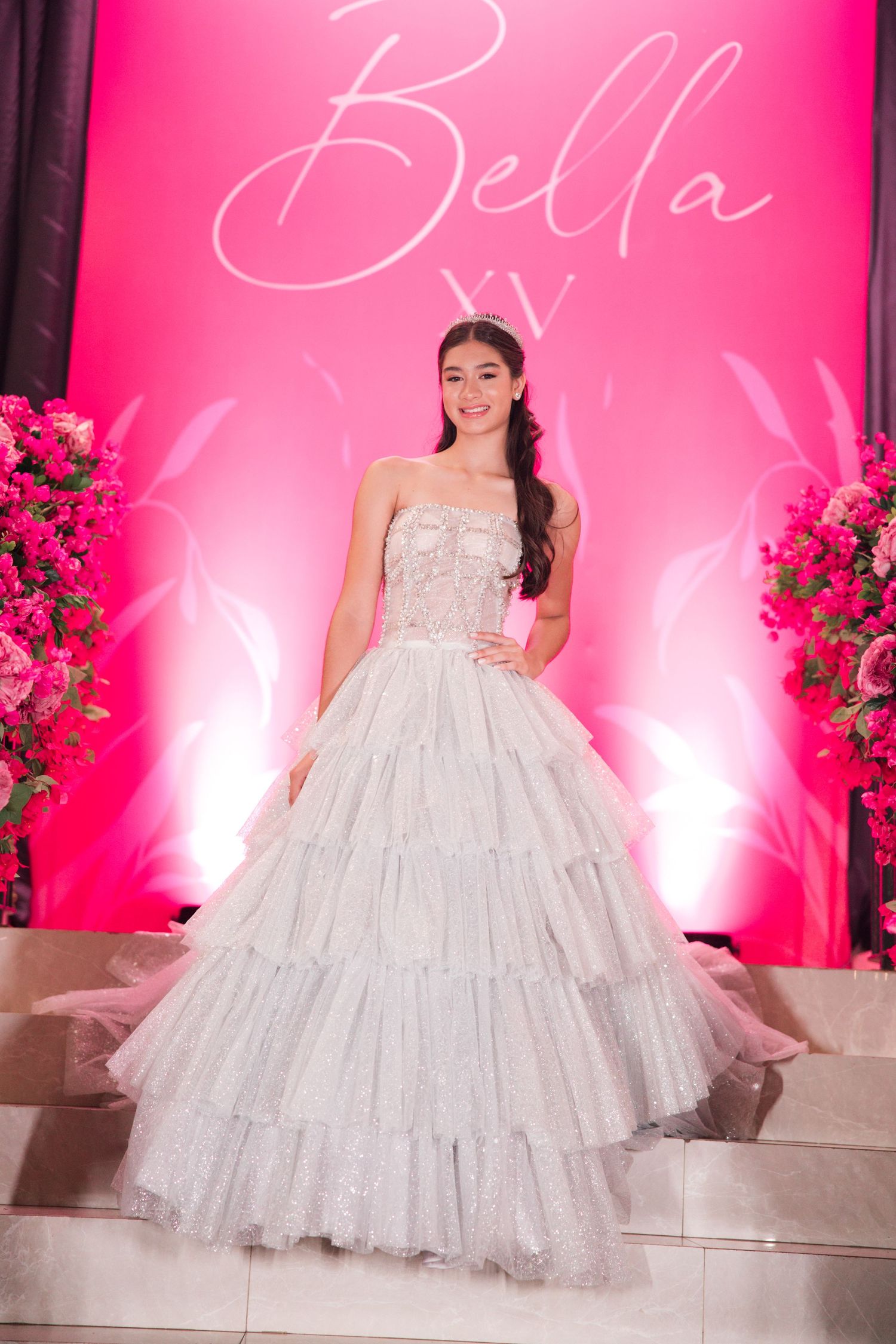 A ballgown-clad person poses in front of a pink backdrop with floral arrangements at a formal event.