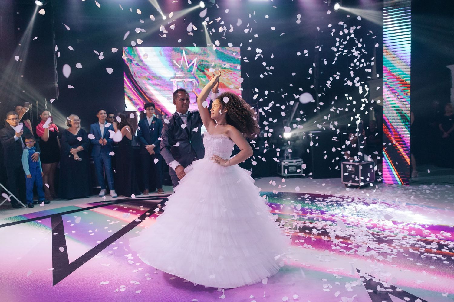 A newlywed couple performs their first dance surrounded by confetti in a beautifully lit reception venue with purple lighting.