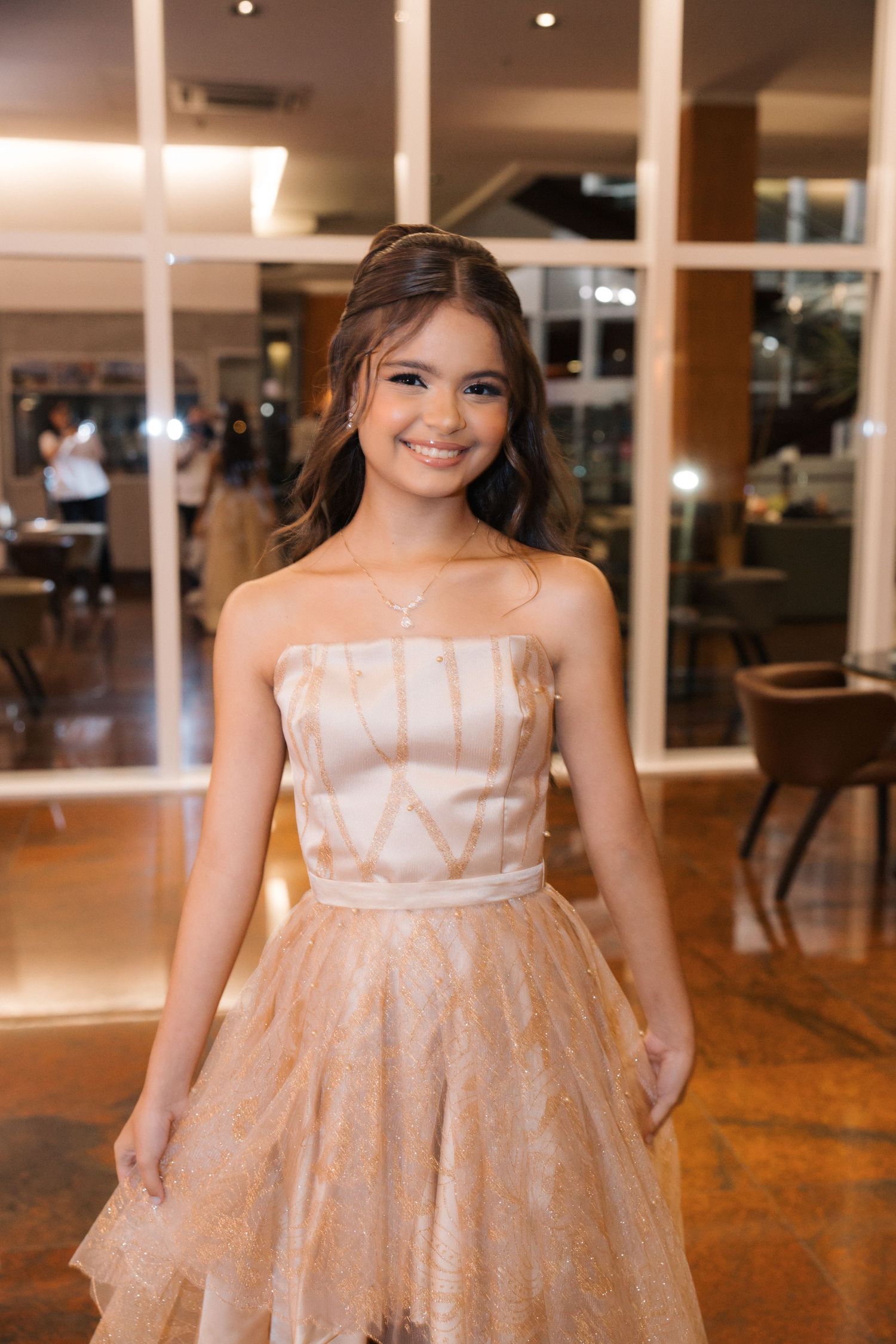 A person in a peach-colored strapless tulle dress stands smiling in an elegant indoor venue with warm lighting.