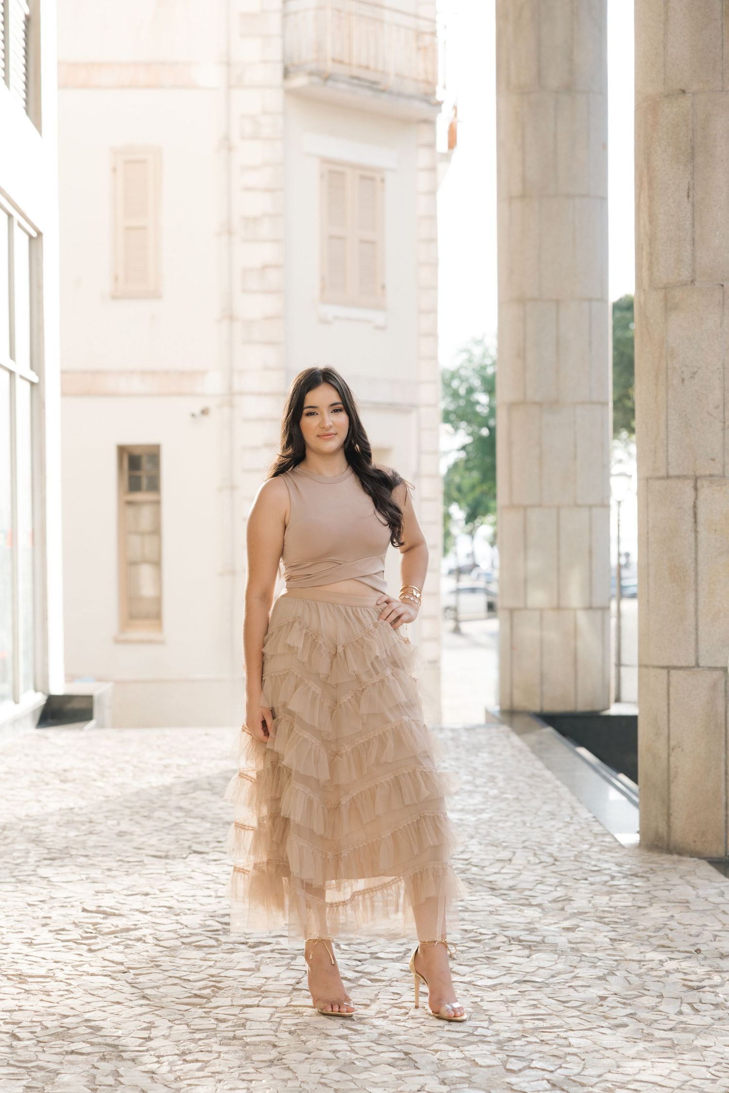 A woman poses elegantly in a beige layered tulle skirt and sleeveless top against a light stone architectural backdrop.