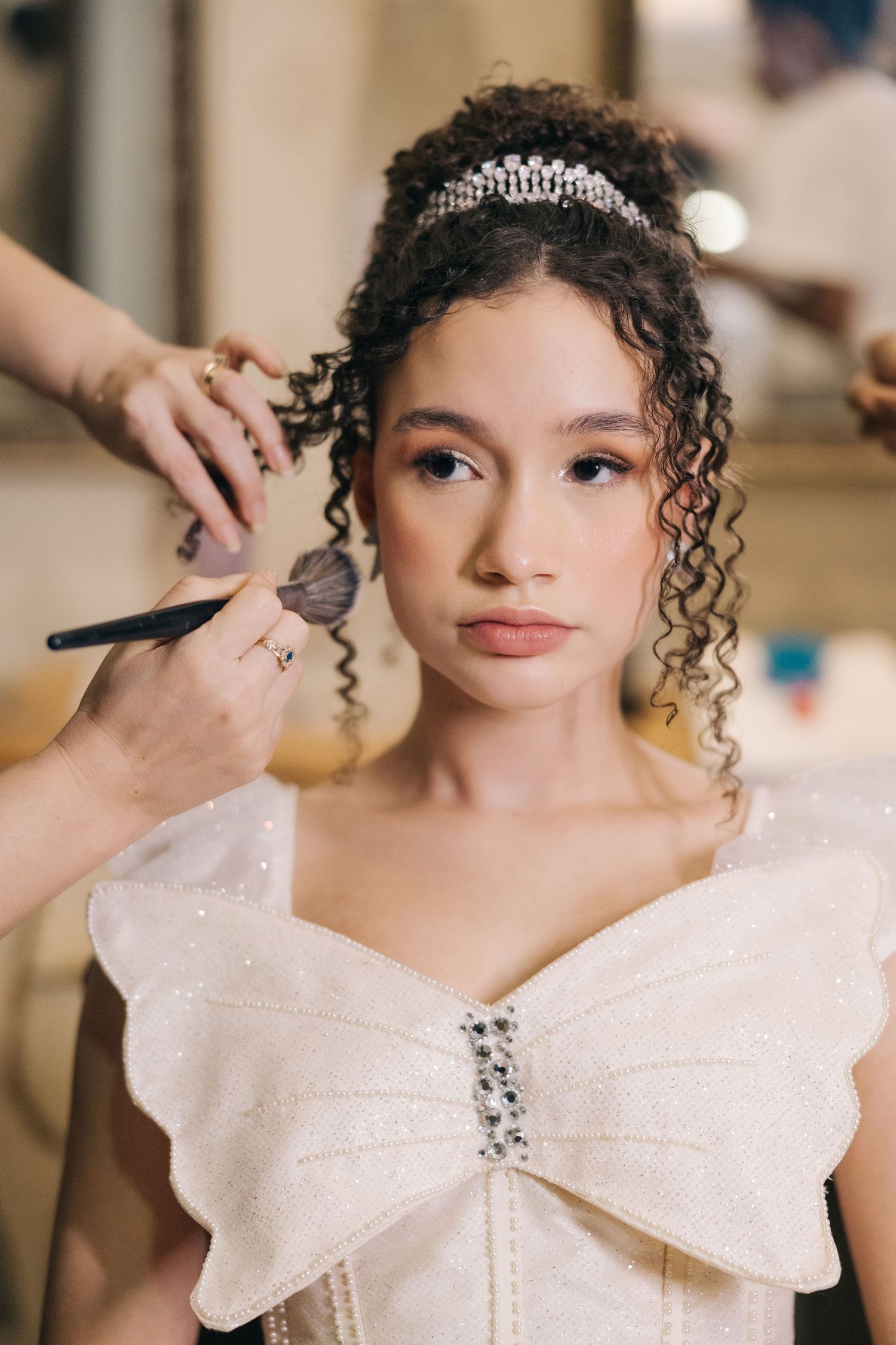 A beautiful wedding day preparation scene shows makeup application on a bride wearing a white bow-detailed dress.