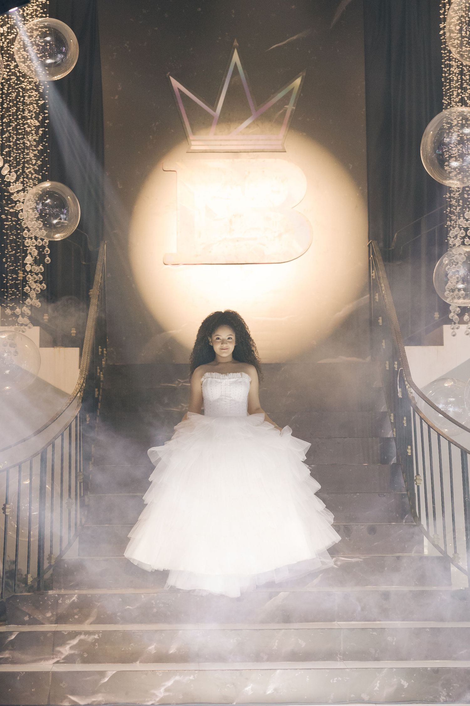 A dreamy portrait on misty stairs featuring a white ballgown under an illuminated crown logo.