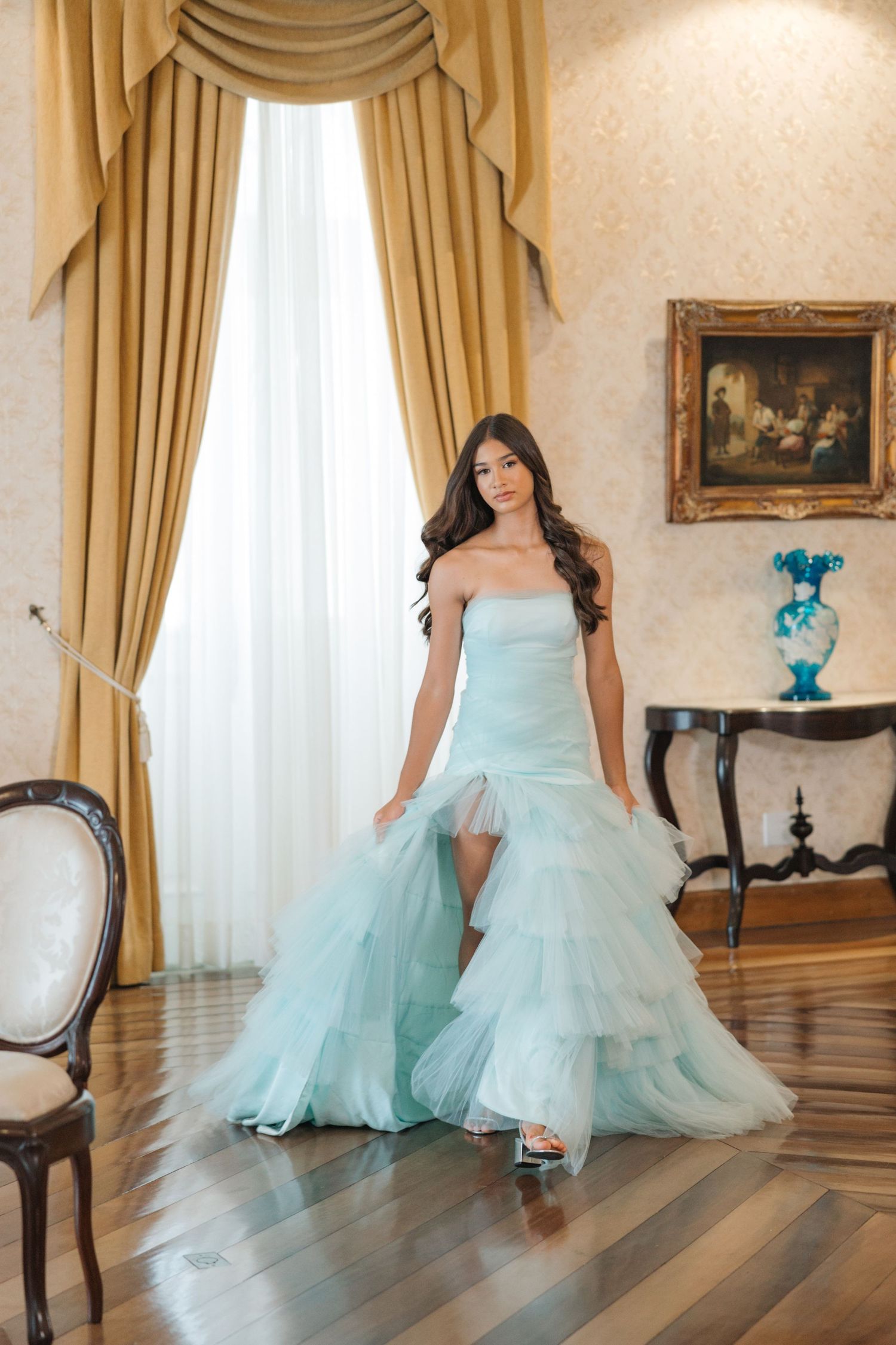 A light blue tulle ball gown with tiered ruffles is showcased in an elegant room with gold curtains and hardwood floors.