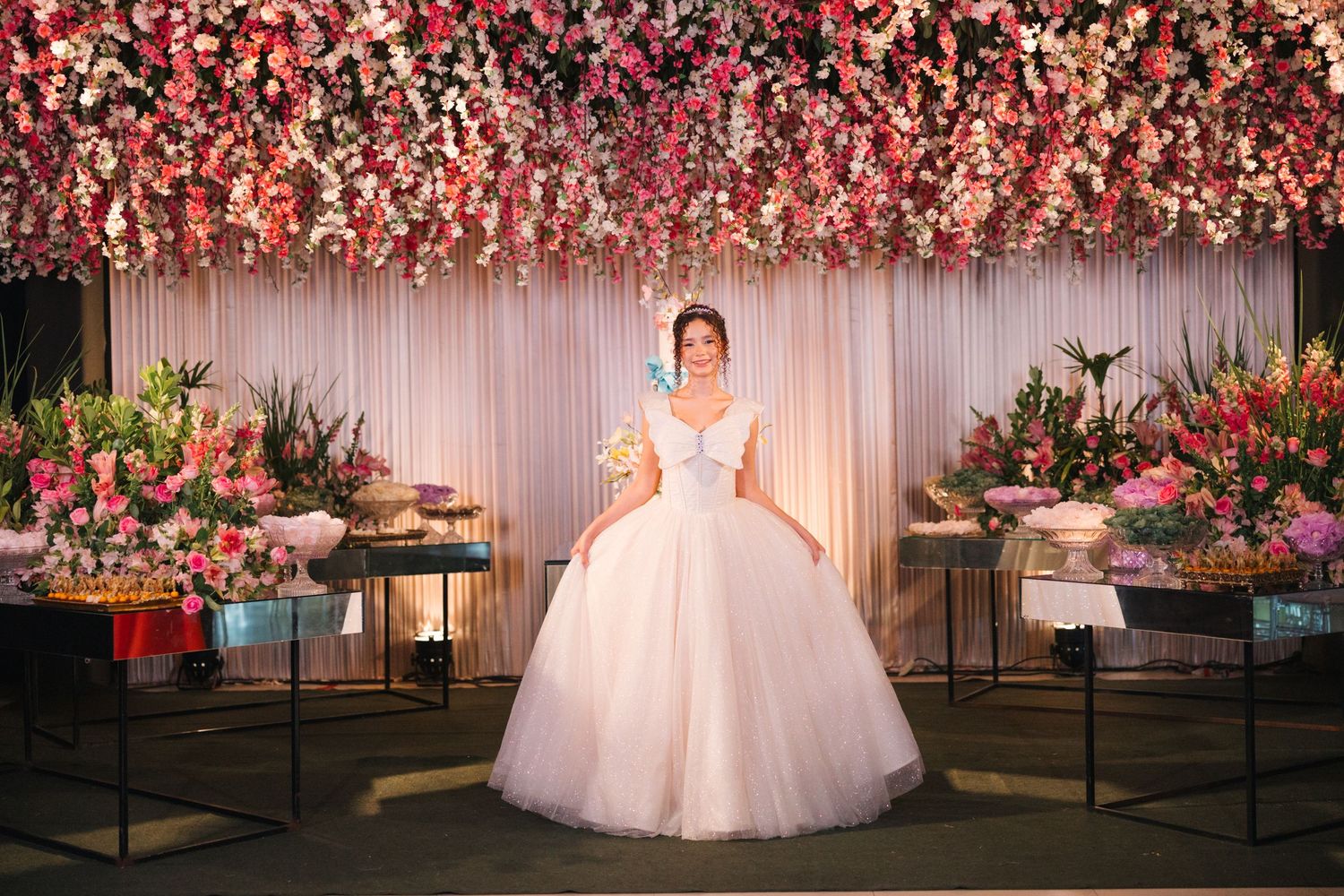 A lavish event space decorated with pink floral arrangements and white drapery features a person in an elegant white ballgown.