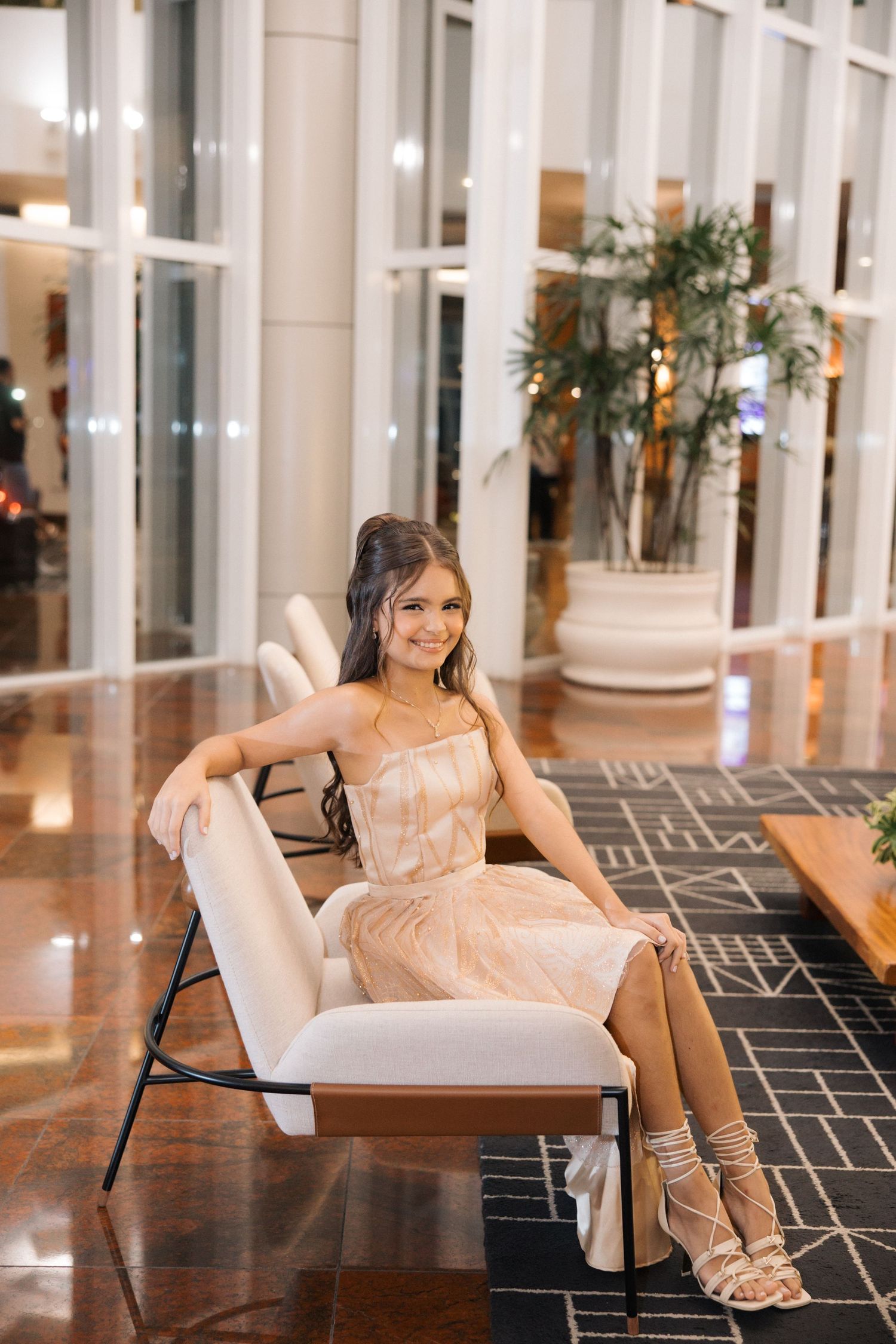 Person in a blush-colored evening dress sits elegantly on a modern white lounge chair in an upscale lobby setting.
