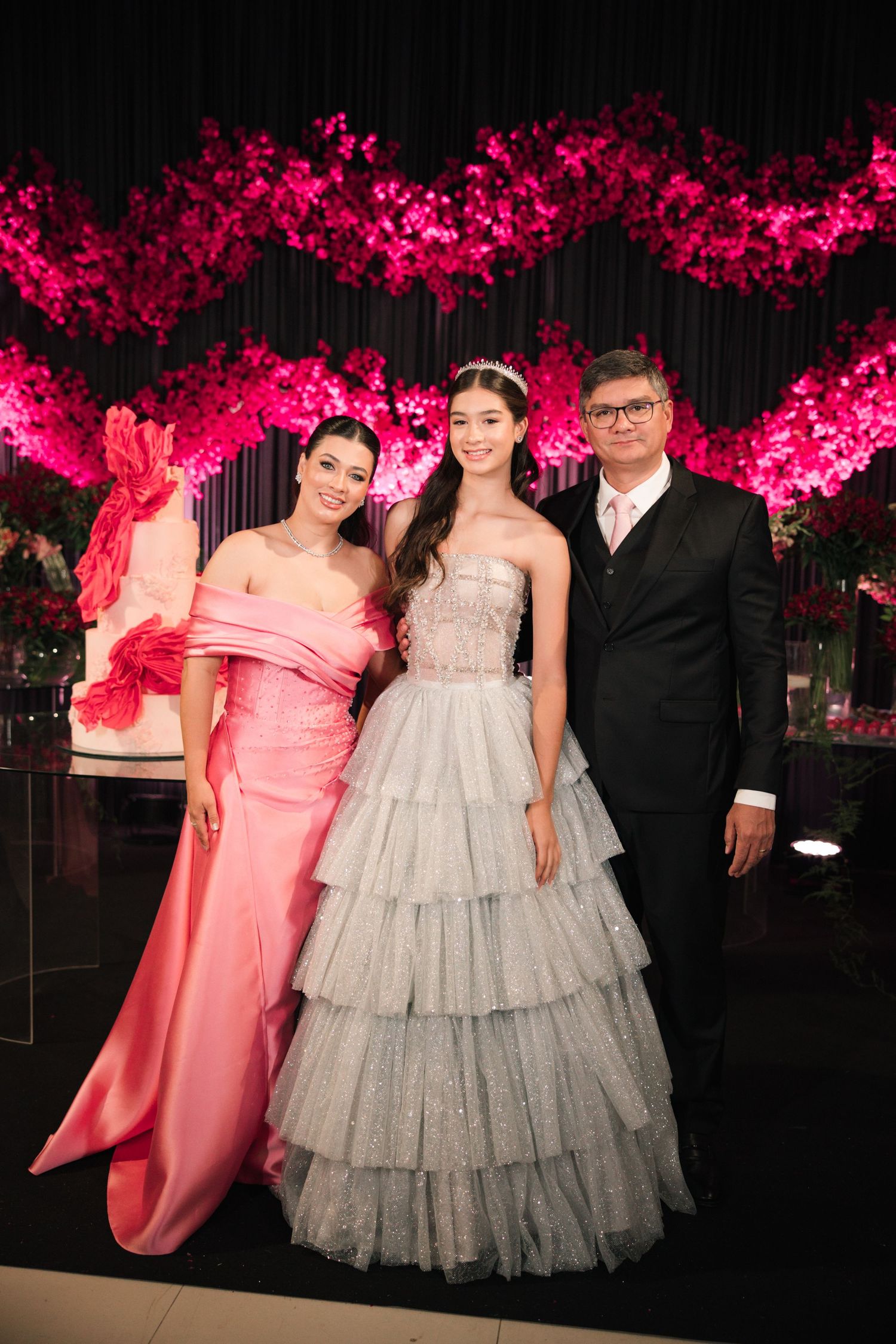 Three people pose at a formal event beneath dramatic pink floral decorations.