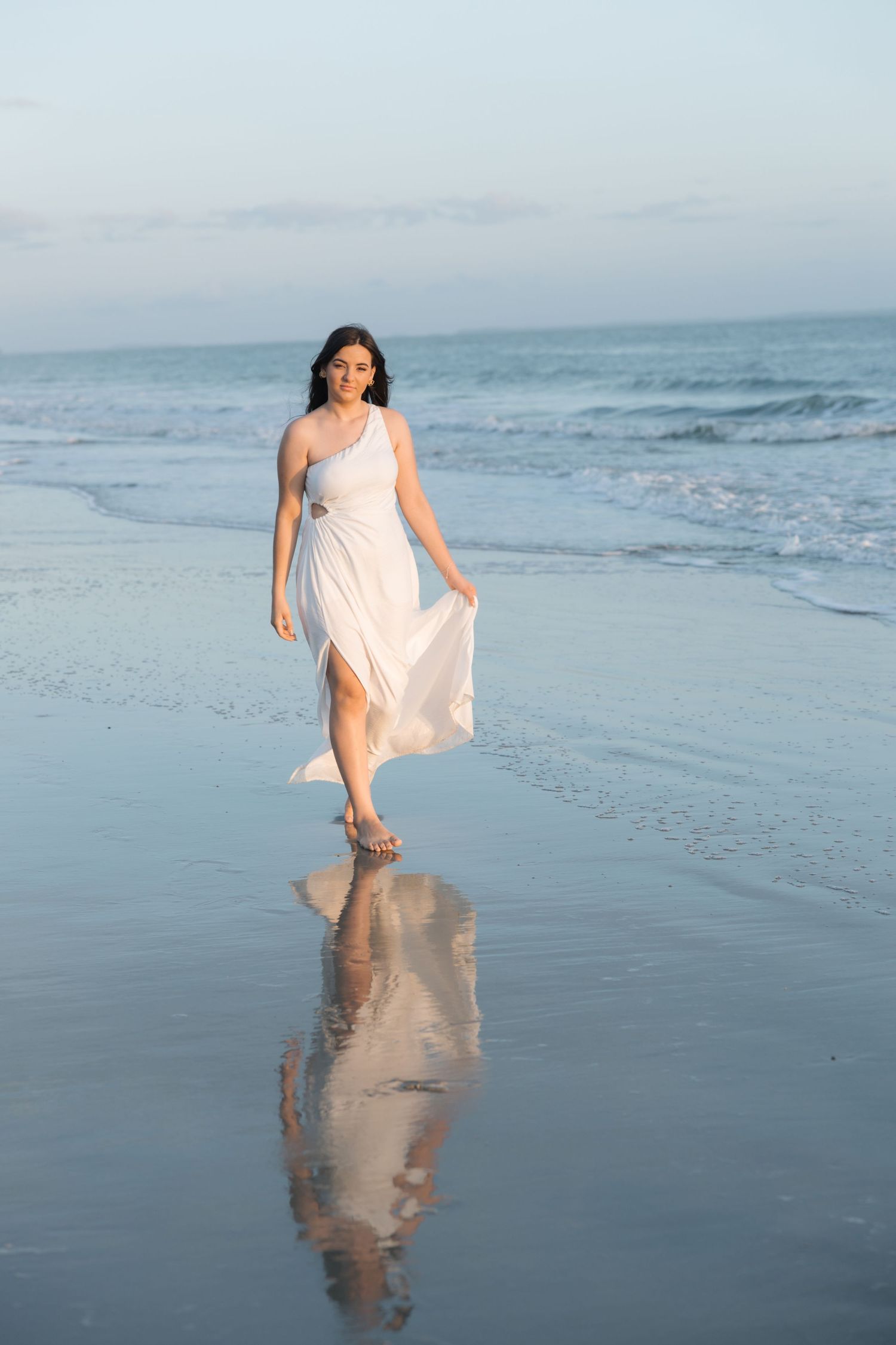 Woman in flowing white dress walks along a beach shoreline at dusk with her reflection visible in the wet sand.