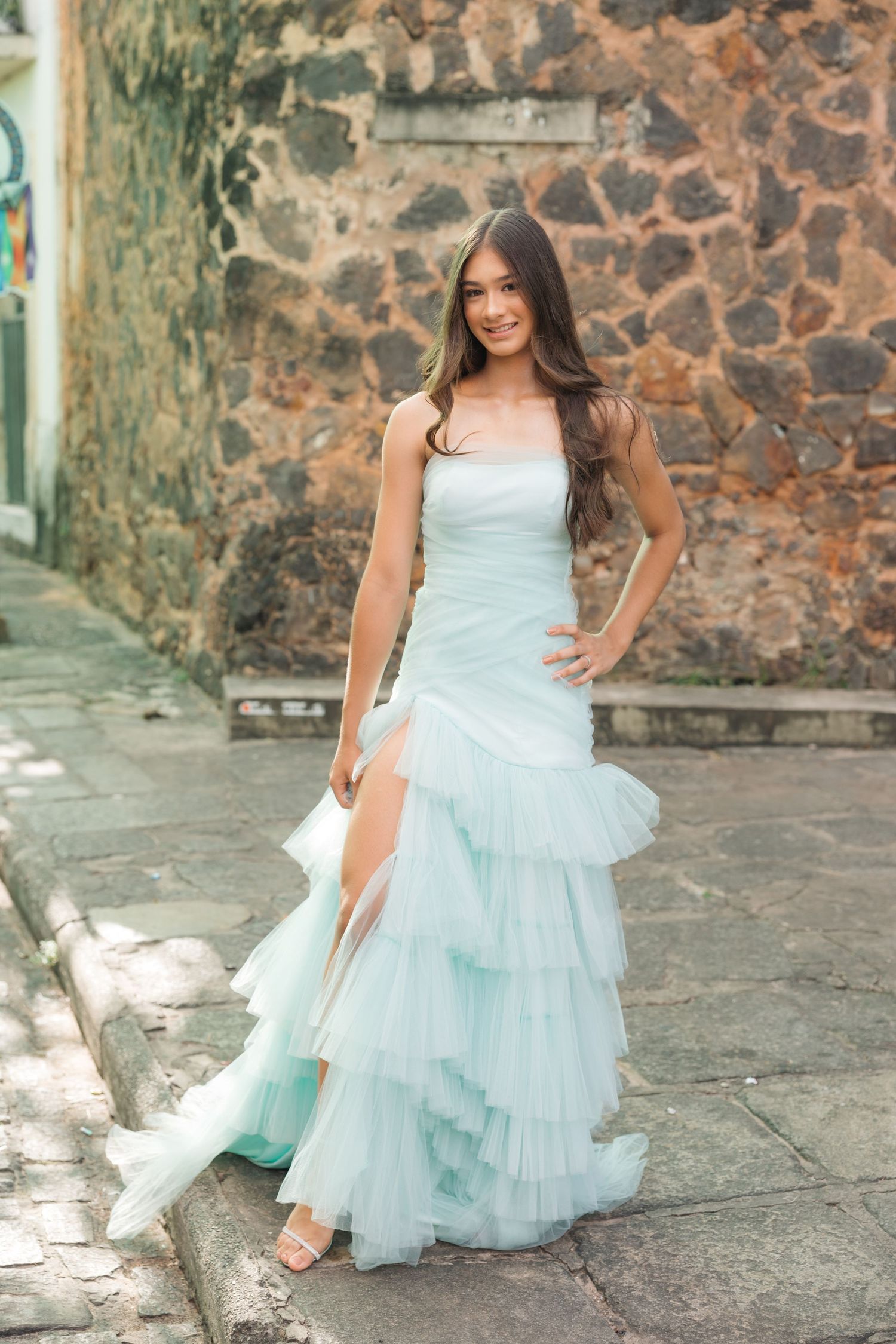 A ruffled light blue tulle gown with a high slit is showcased against a rustic stone wall backdrop.
