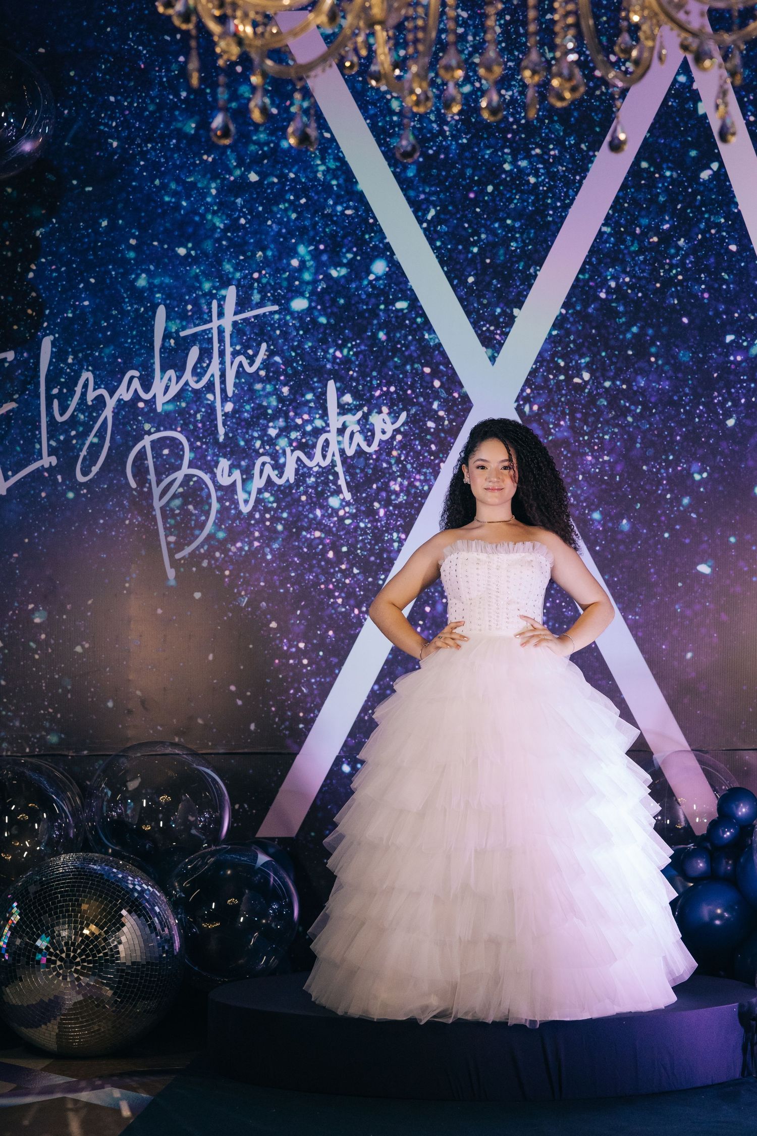 A debutante poses in a white tulle ballgown under a sparkling chandelier with a starry night backdrop.