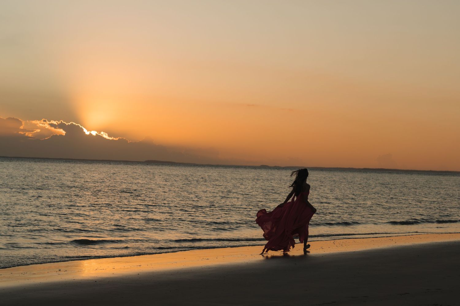 A silhouette walks along the beach at sunset with flowing dress trailing behind.