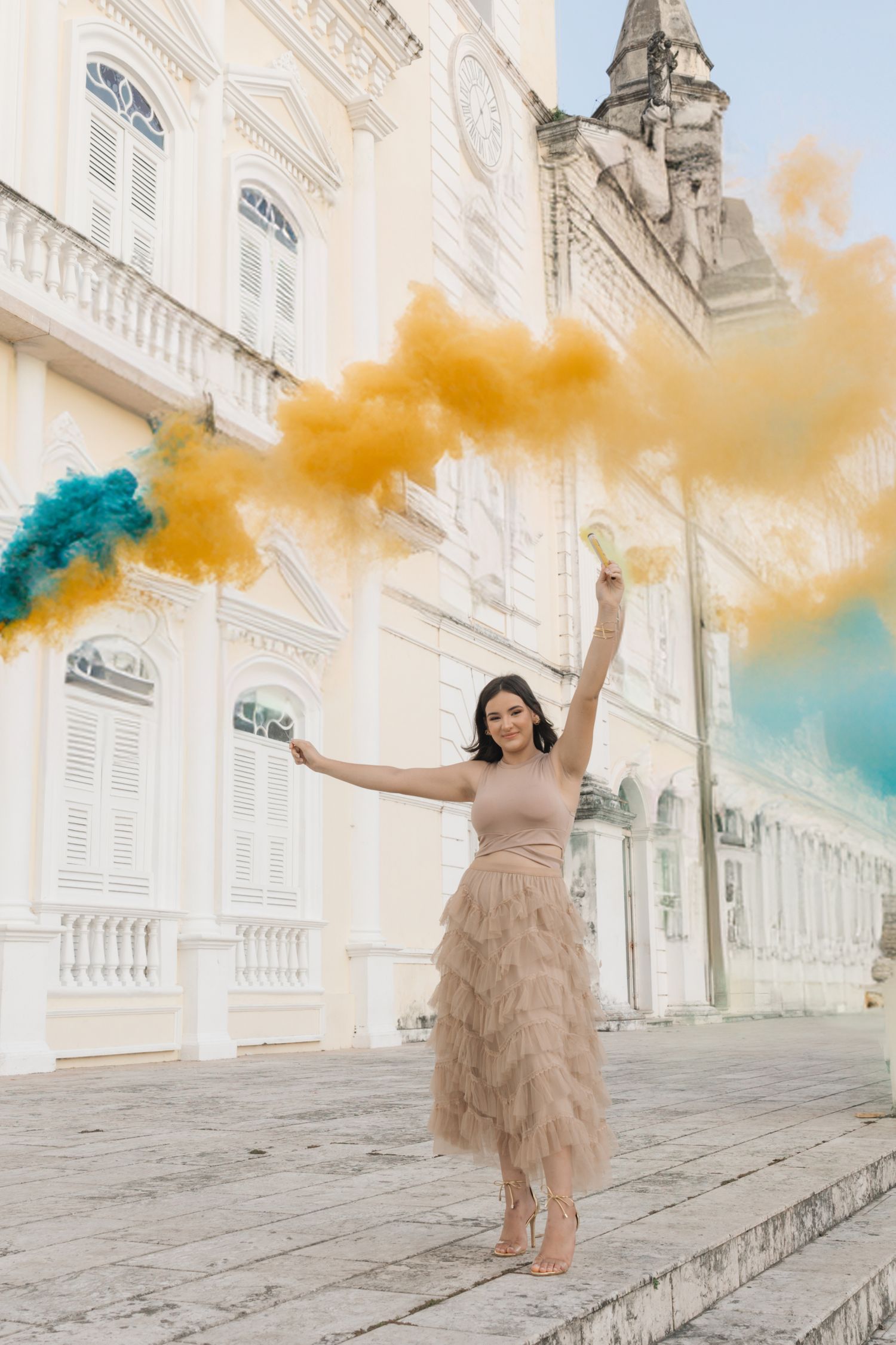 Woman in beige ruffled dress poses with colorful smoke bombs outside ornate white classical building.