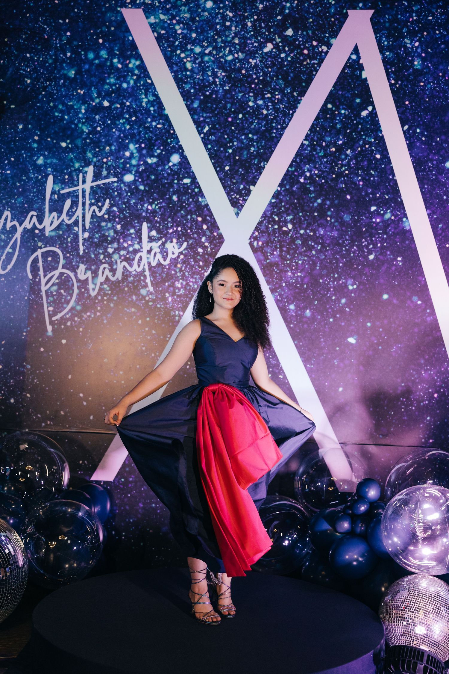 Woman in navy dress with red underskirt poses against starry backdrop with large illuminated letter display.