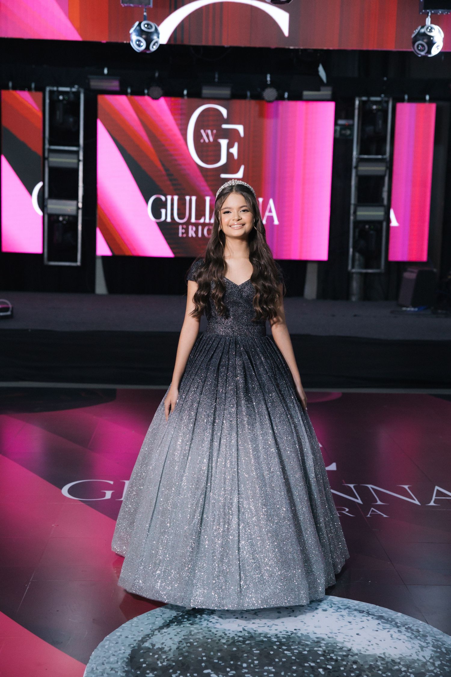 A dramatic ball gown on display at a Giuliana fashion event with pink and red backdrop lighting.