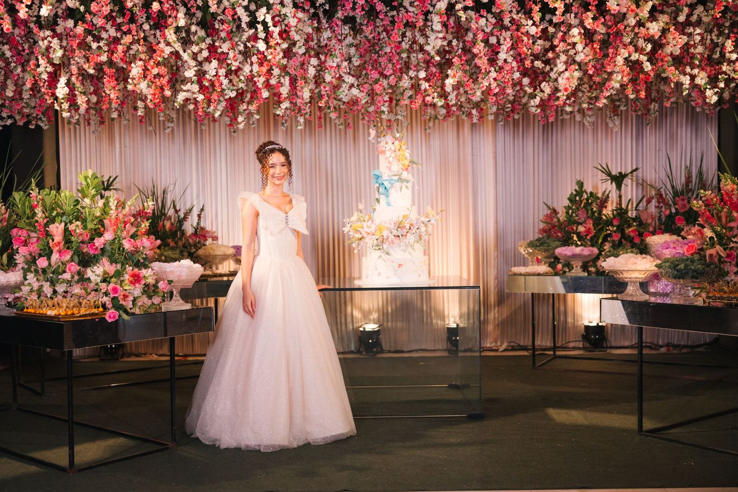 Elegant wedding reception setup with pink floral canopy, white tulle gown, illuminated backdrop and dessert display tables.