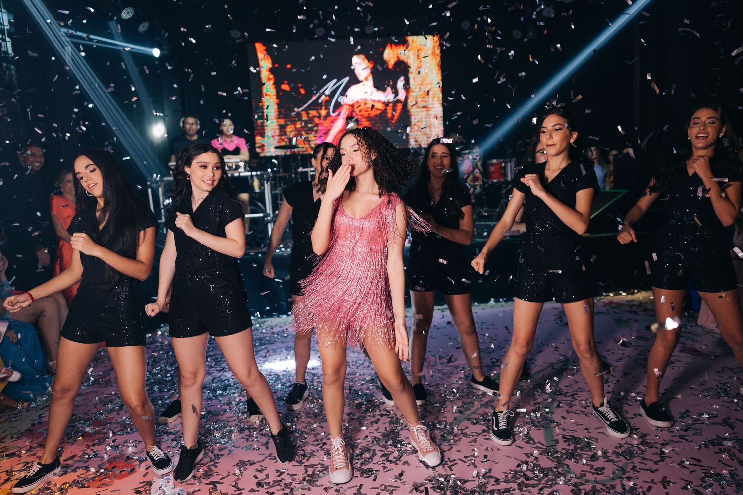 Group performs choreographed dance routine in black outfits with pink stage lighting and confetti.