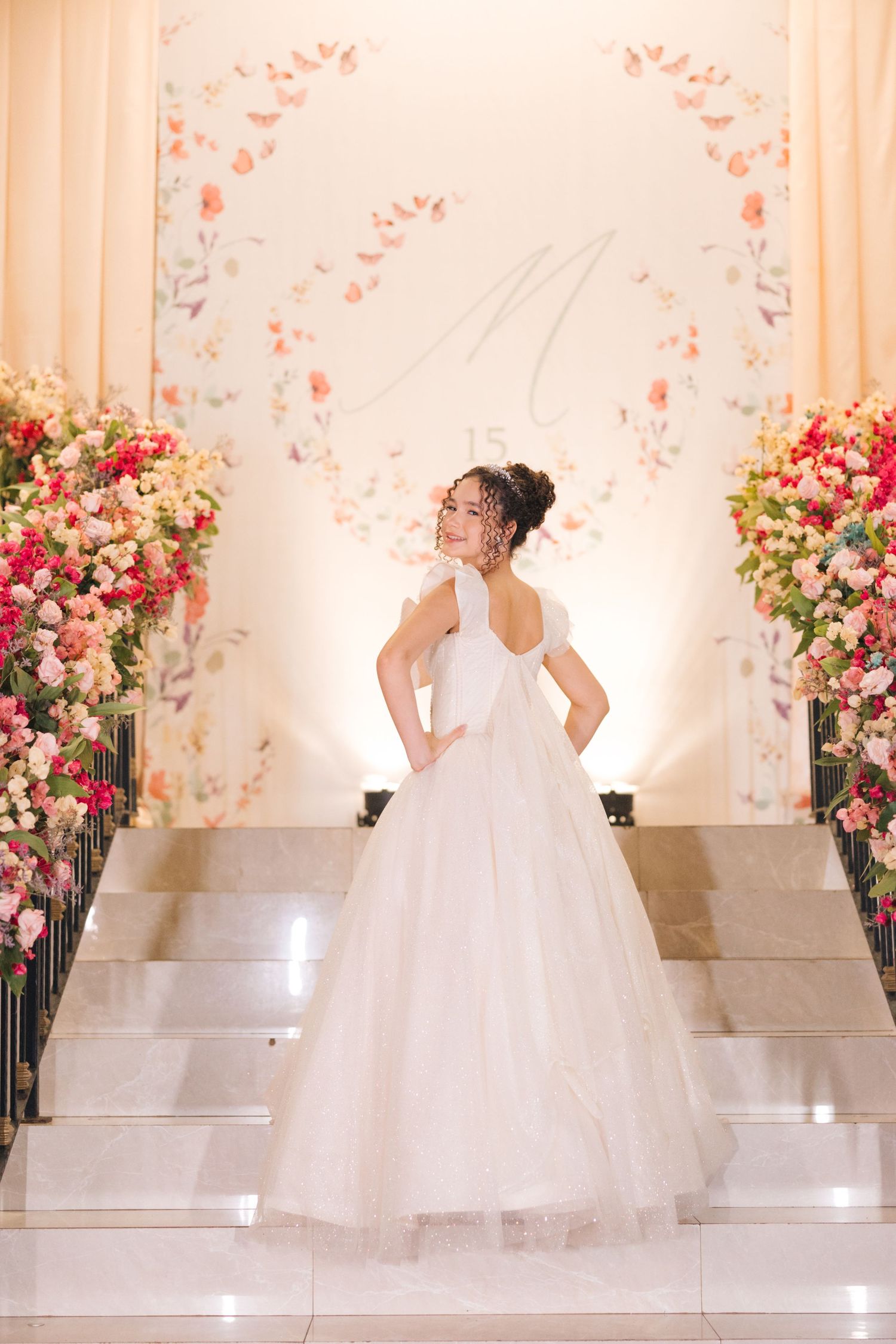 Back view of a white wedding gown with ruffle details and tulle skirt against a floral backdrop.