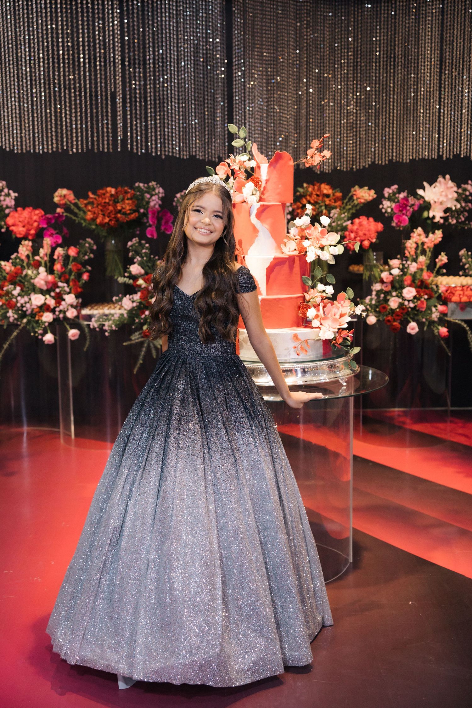 A debutante in a sparkling ombre gown stands next to a coral-colored wedding cake surrounded by floral decorations.
