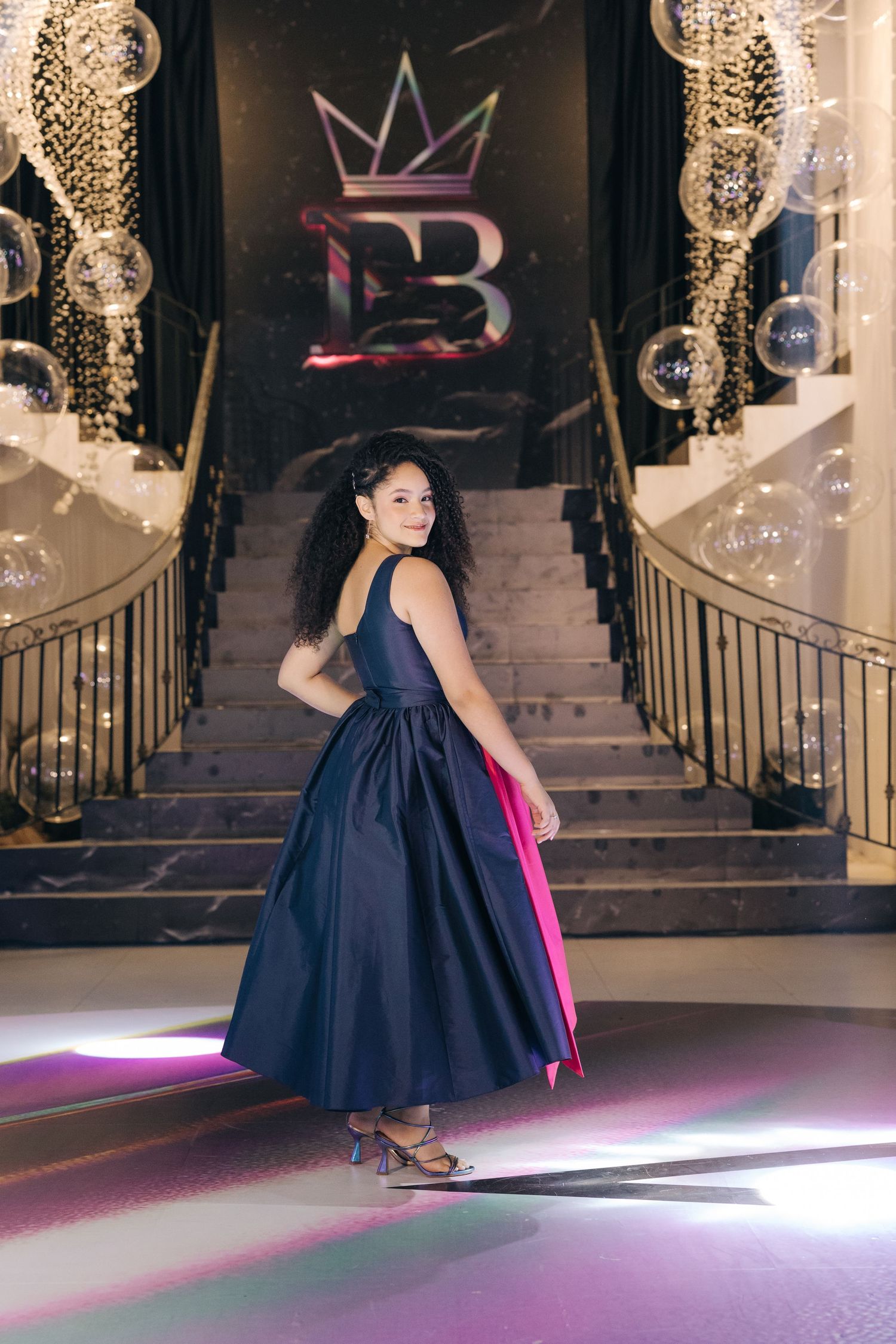 Elegant woman in navy blue ball gown poses on grand staircase with illuminated crown logo and decorated railings.
