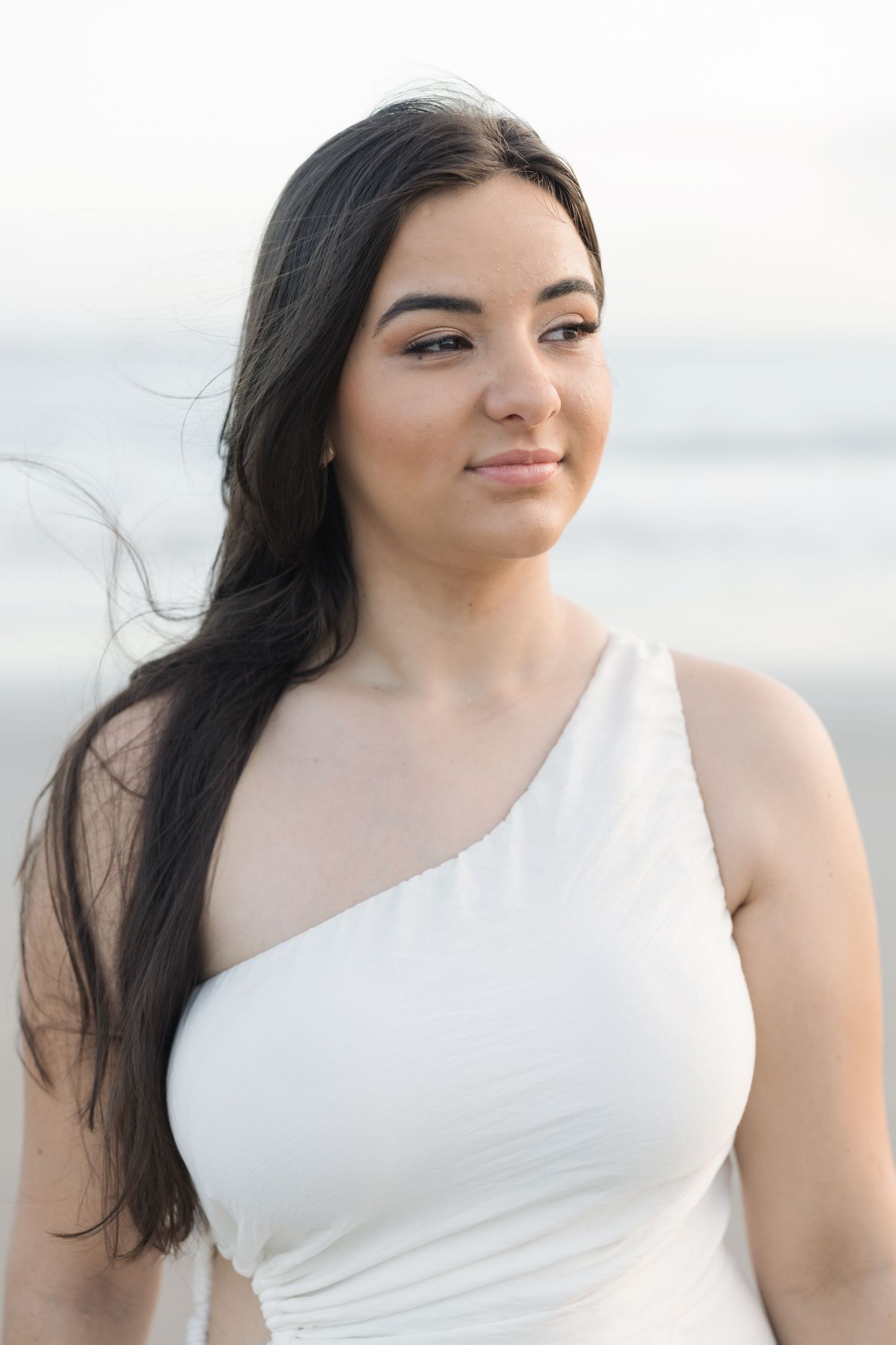 Portrait in white one-shoulder dress with long dark hair against a soft, bright background outdoors.