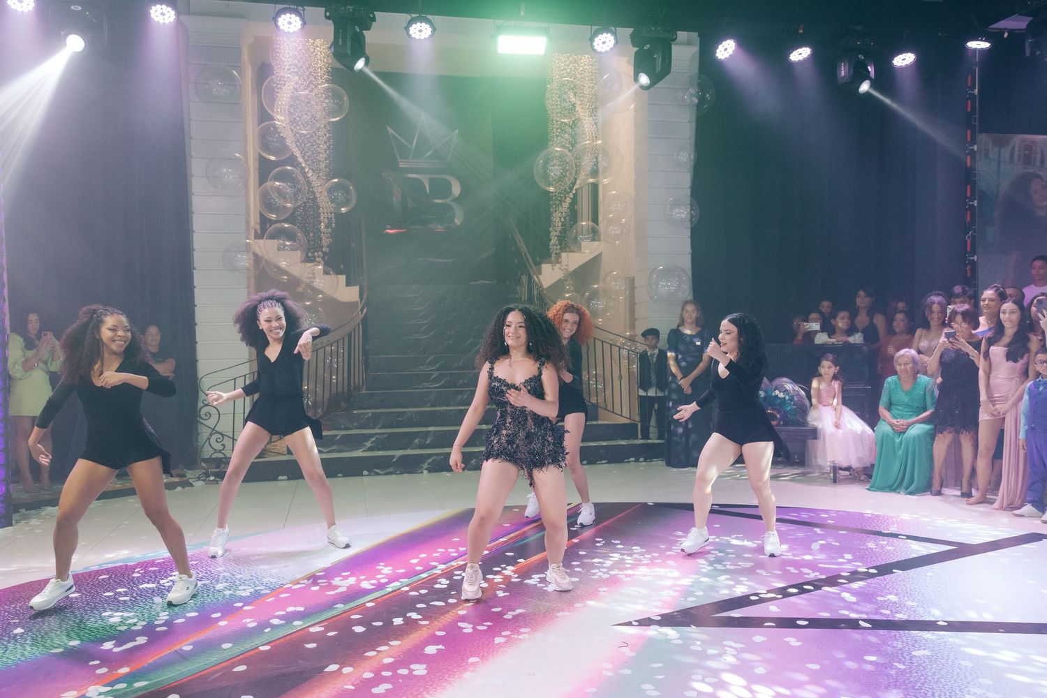 Dancers perform on stage with colorful lights and confetti while audience members watch from the sides.
