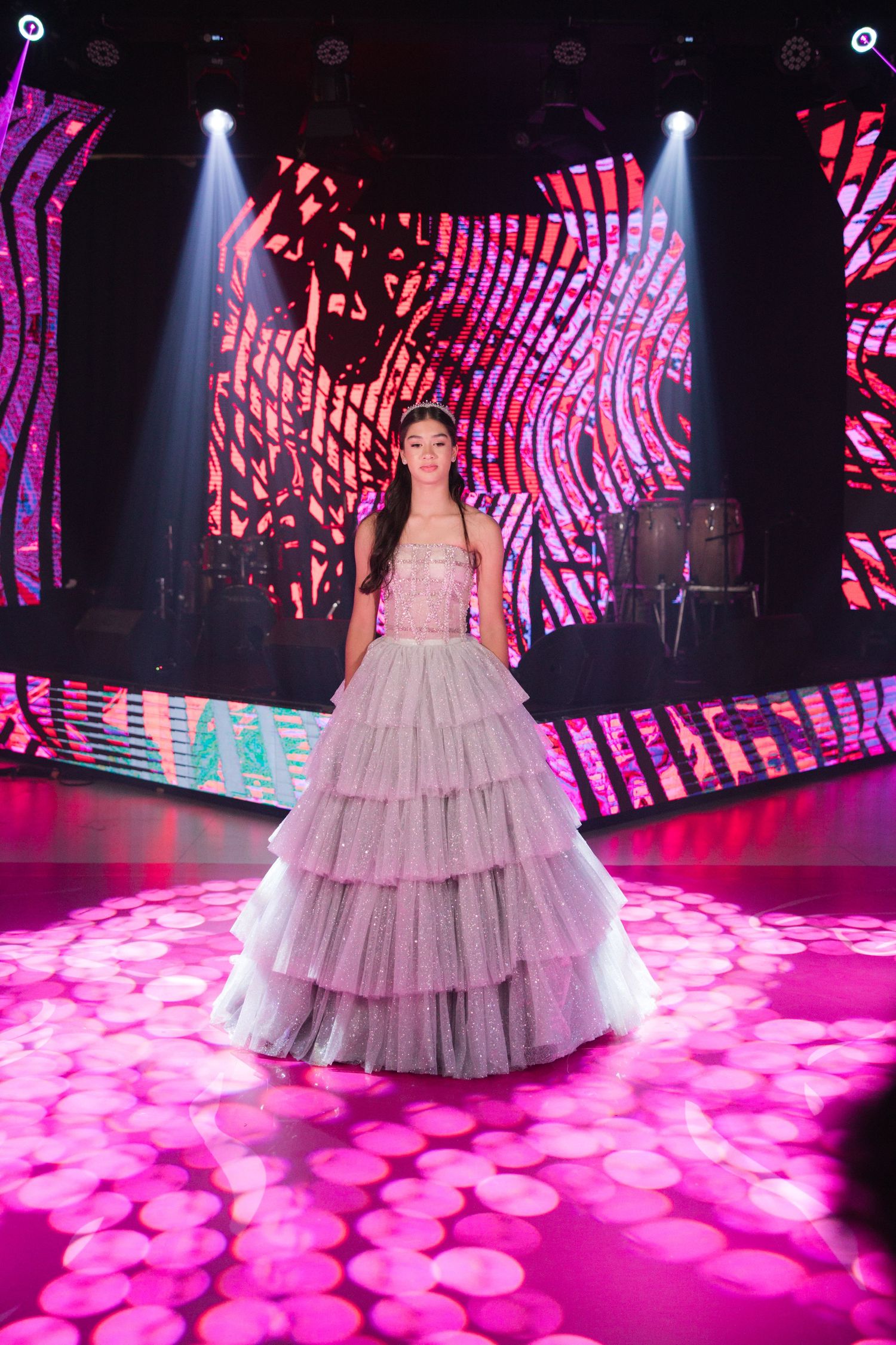 A flowing tiered ballgown captured under dramatic pink stage lighting with geometric LED displays.
