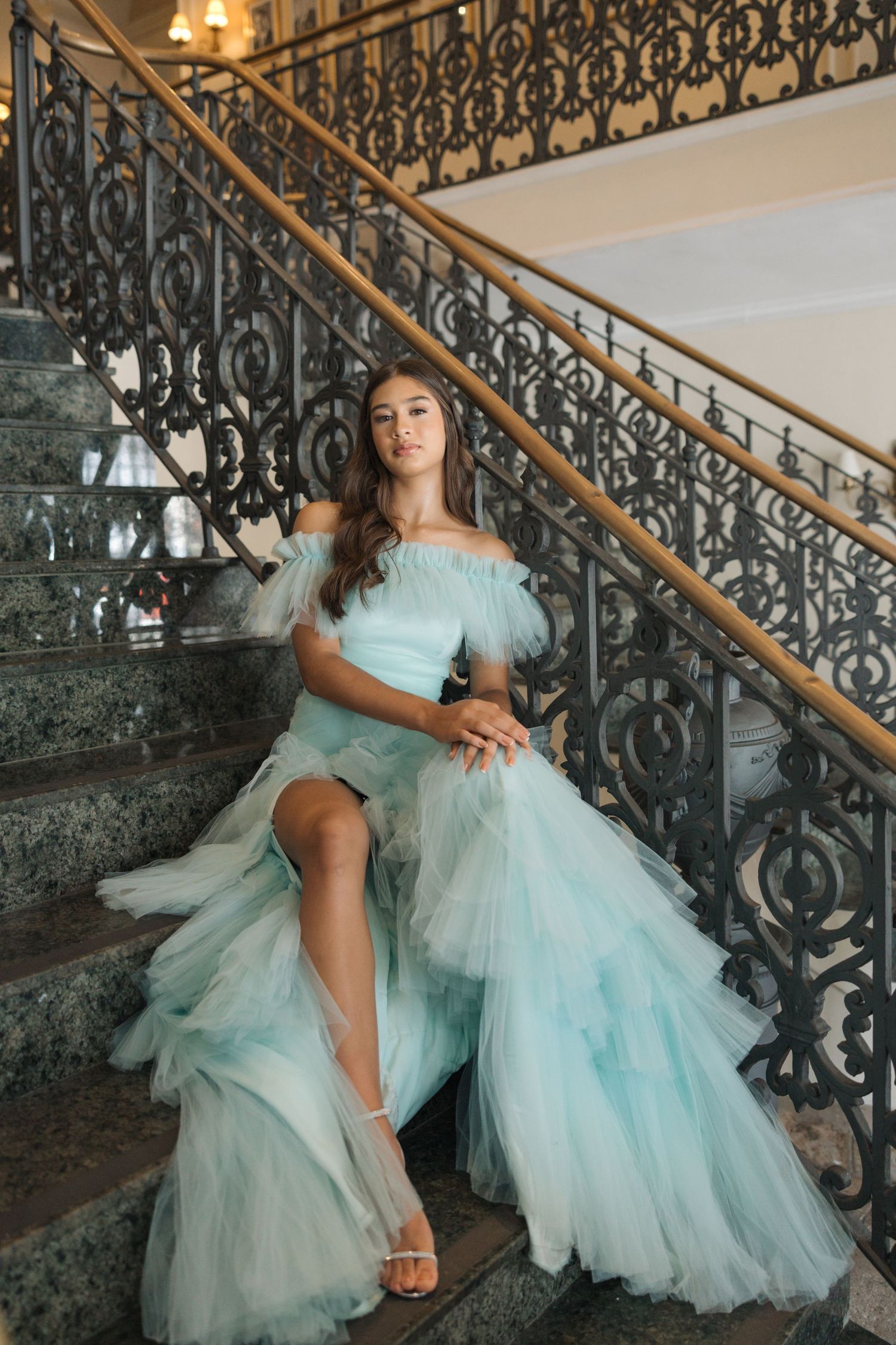 An elegant off-shoulder mint tulle gown flows gracefully along ornate wrought iron staircase railings.