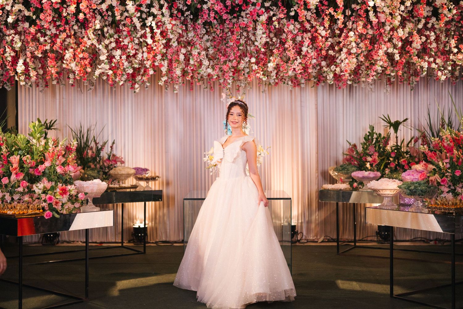 A person stands in a white gown against a floral wall backdrop with pink blooms and elegant side table decor.