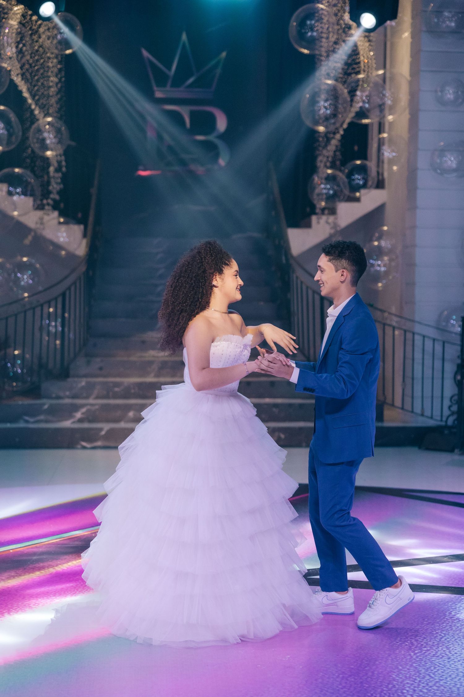 Couple sharing romantic moment at formal event with elegant staircase and crown logo backdrop.