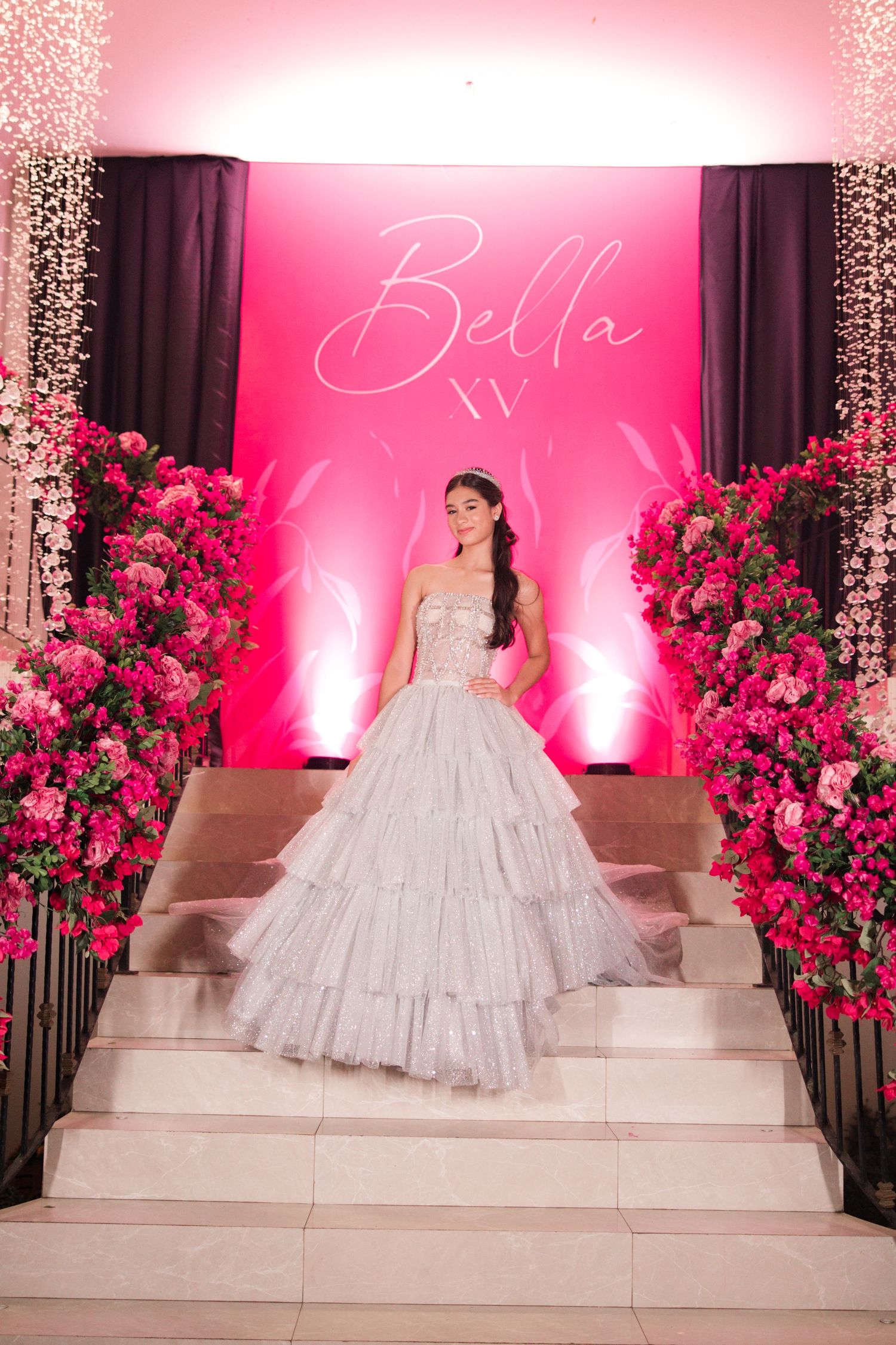 A grand quinceañera celebration with a pink backdrop, floral arrangements and a tiered ballgown on elegant marble stairs.
