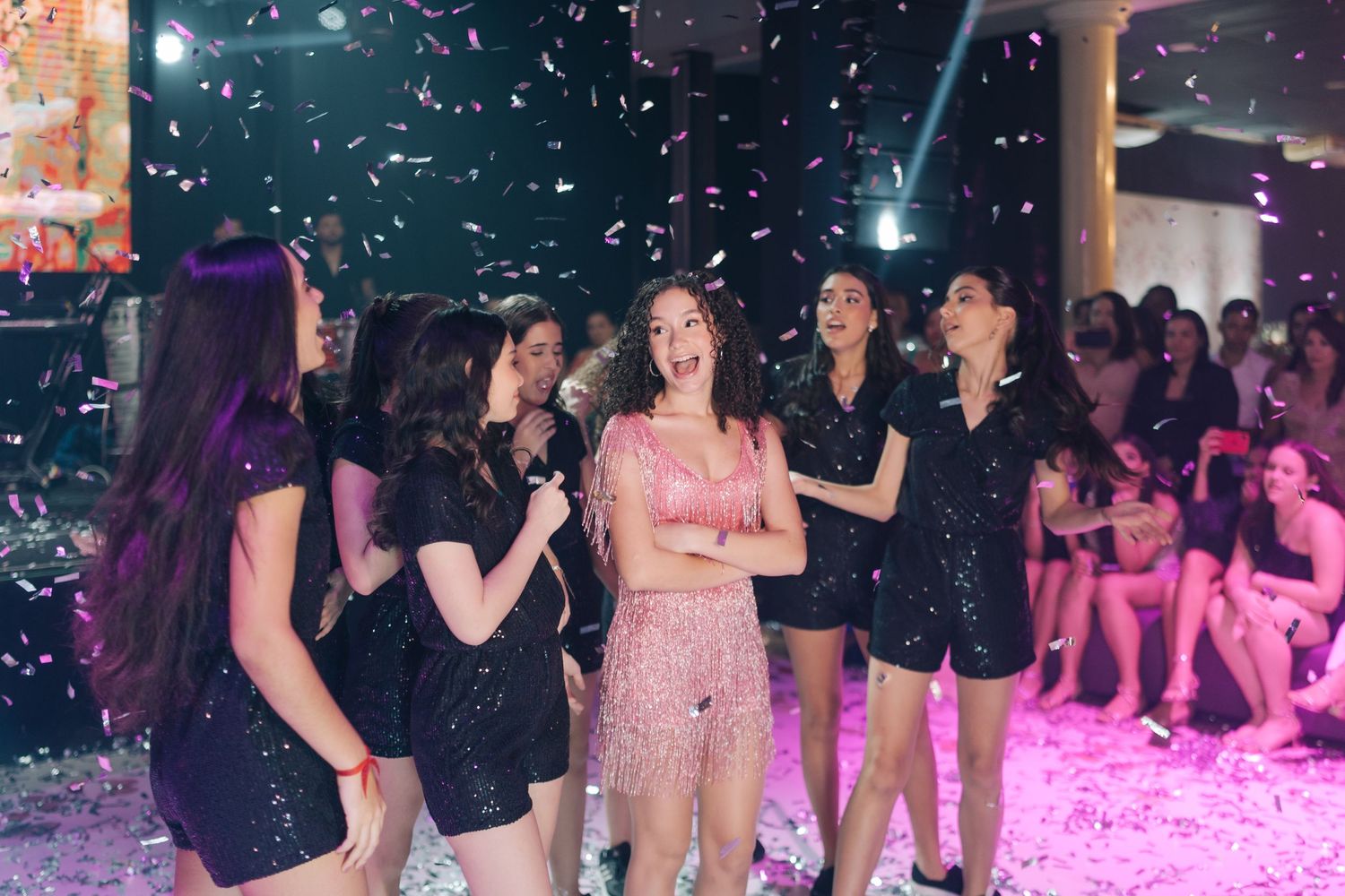 Party celebration with confetti falling and people dancing under colorful lighting.