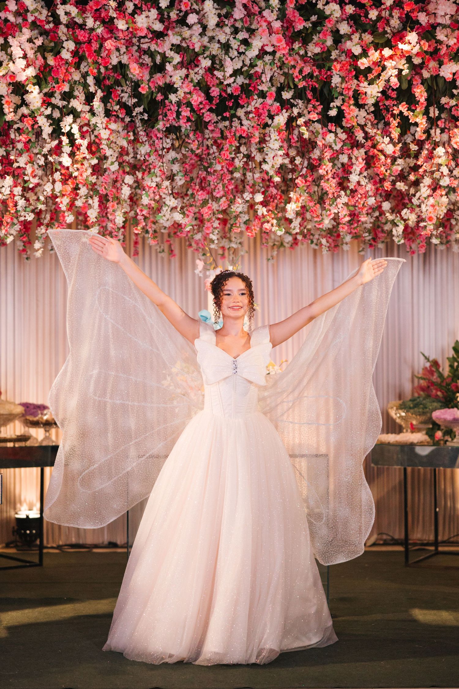 A bride in a flowing white tulle gown poses under a floral canopy with pink and white blooms.