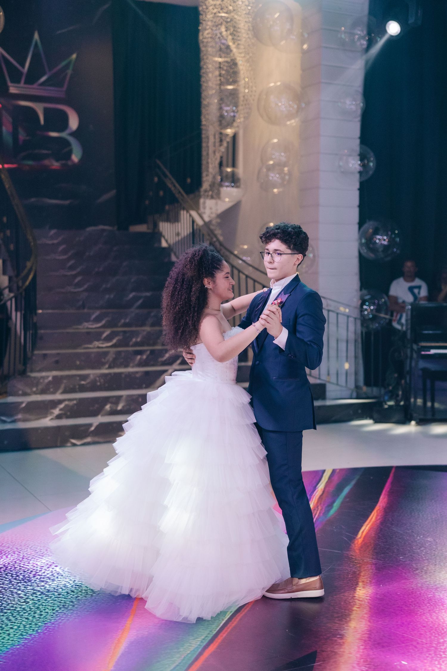 A couple dances together on a colorful illuminated floor beneath a grand chandelier and crown logo.