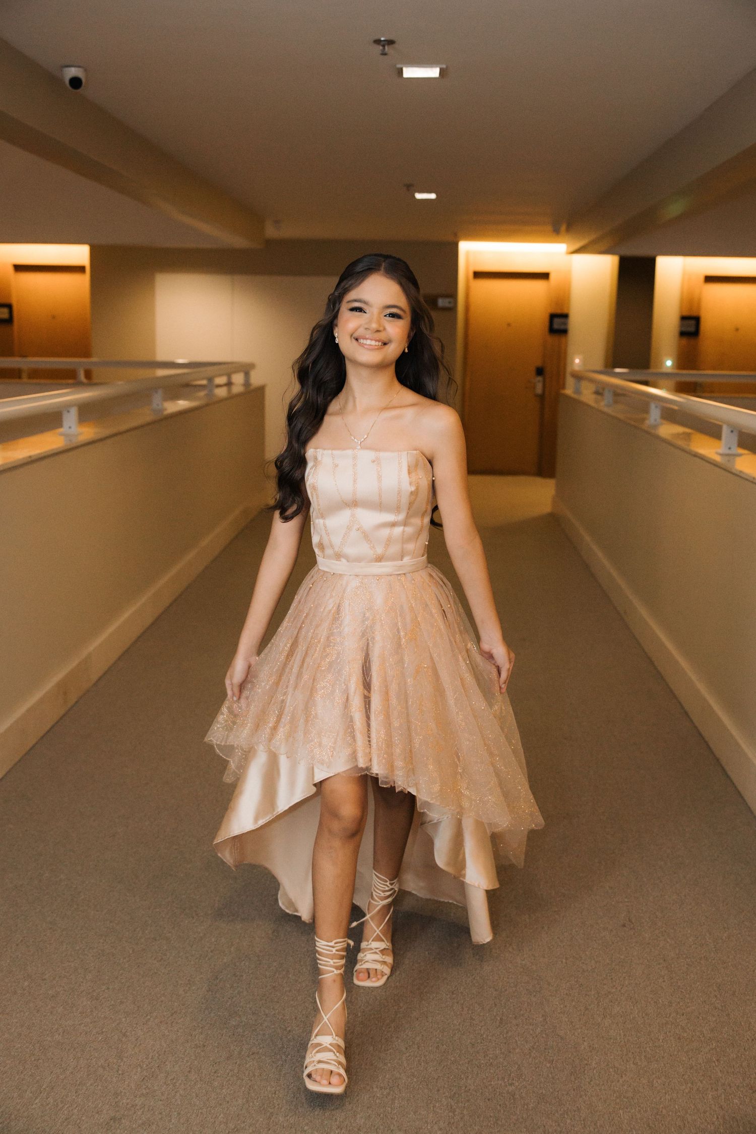 A peach-colored high-low formal dress with sparkles shimmers in a beige hallway interior.