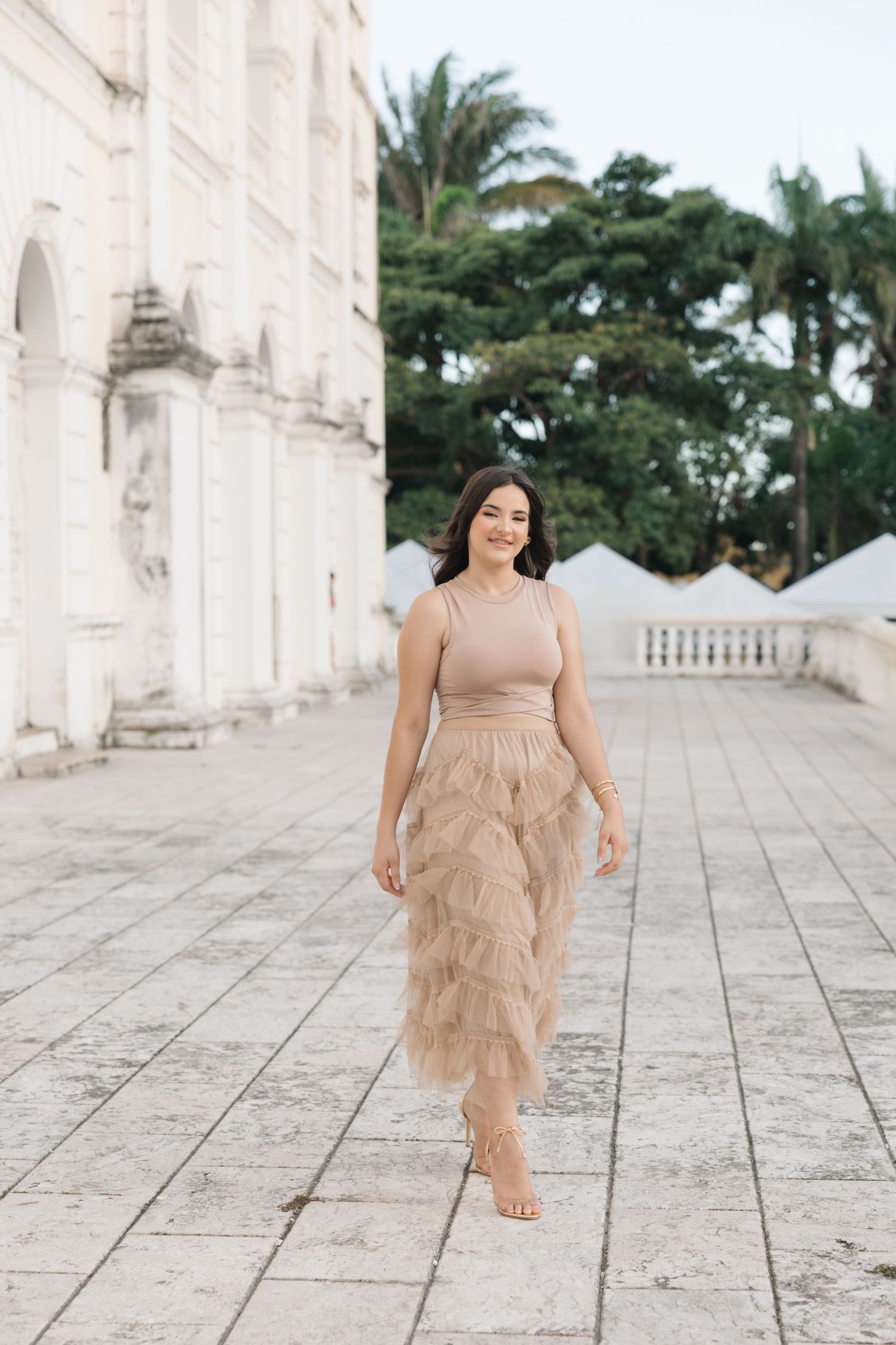 Woman in nude-colored tiered dress walks barefoot along stone pathway at elegant white building with palm trees.