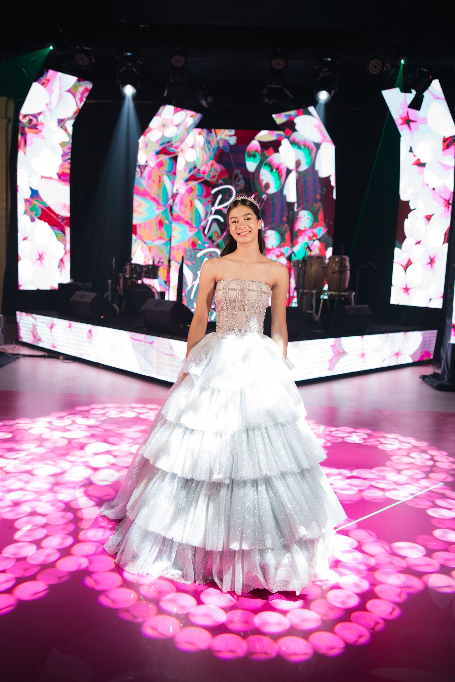 Sparkling silver tiered gown captured on pink-lit dance floor with digital screens and spotlights.