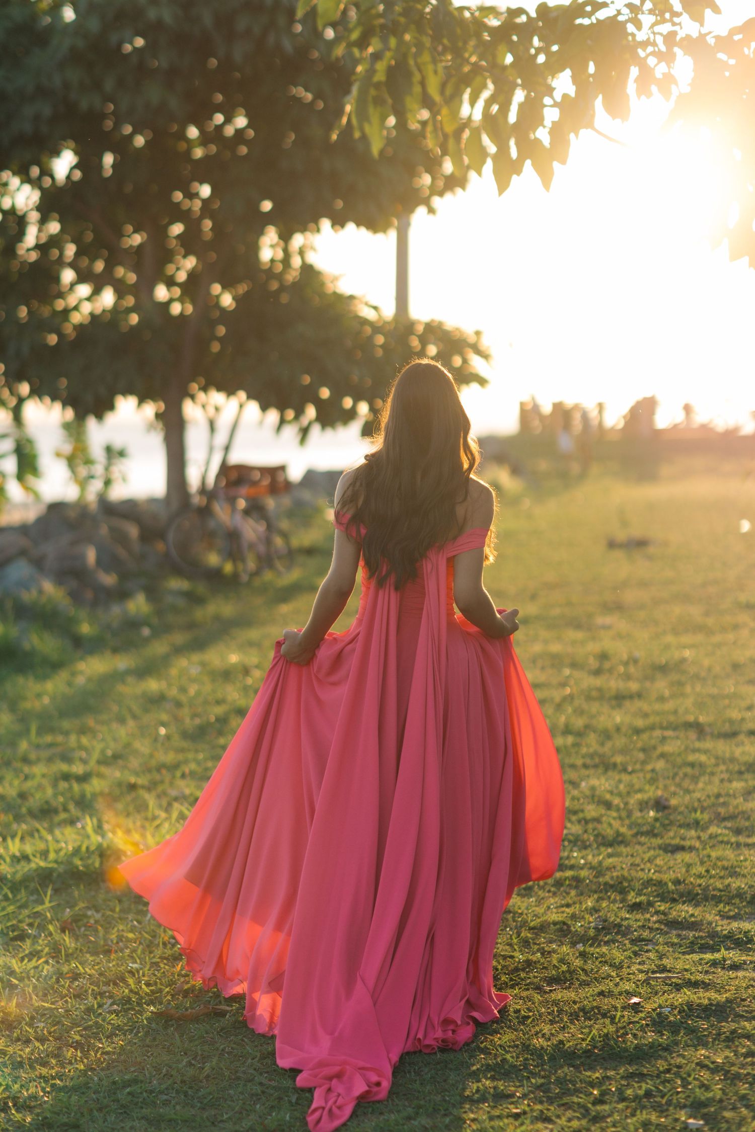 A flowing coral dress silhouetted against a golden sunset in a grassy outdoor setting.