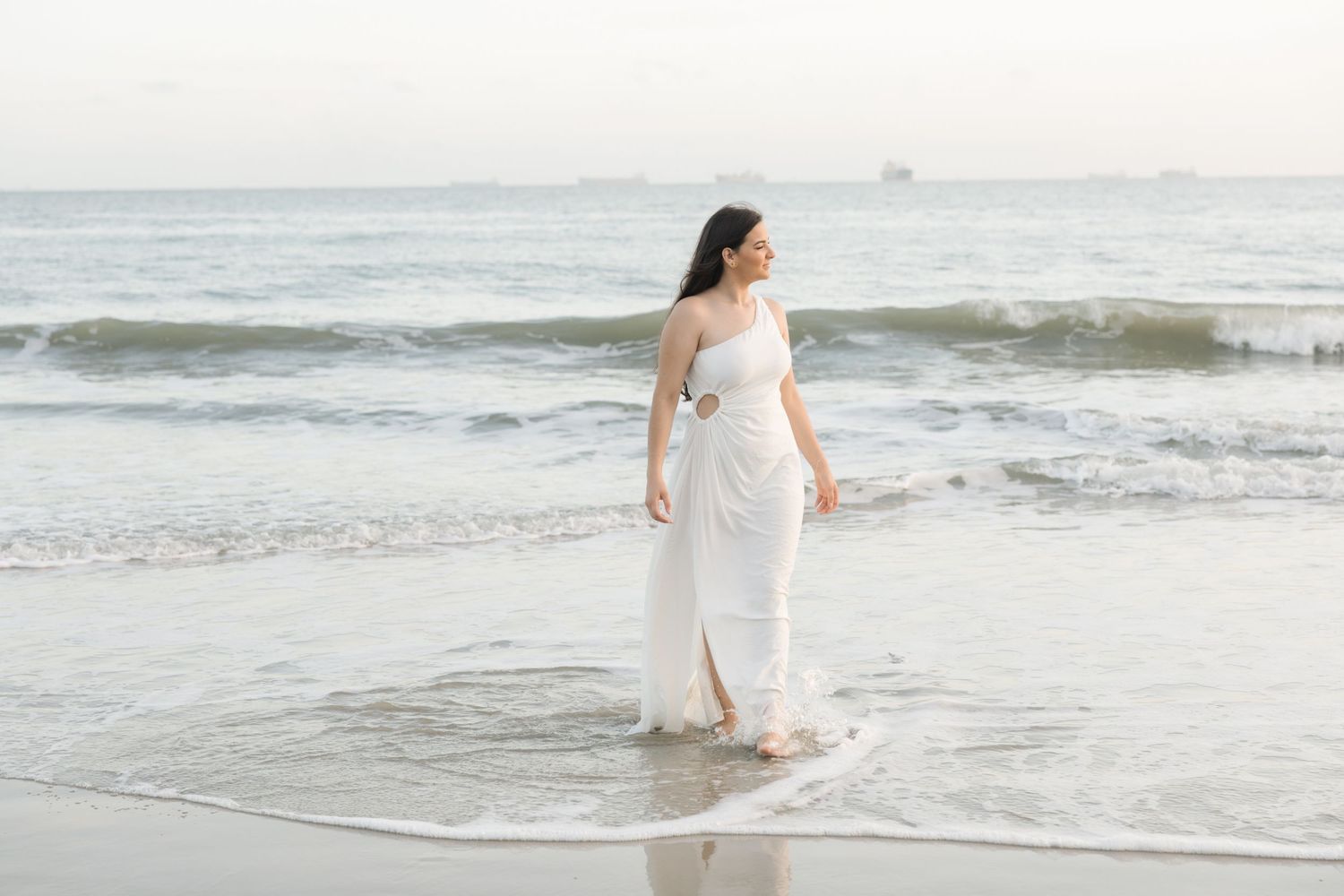 Bride in flowing white dress walks along ocean shore during sunset beach photoshoot.