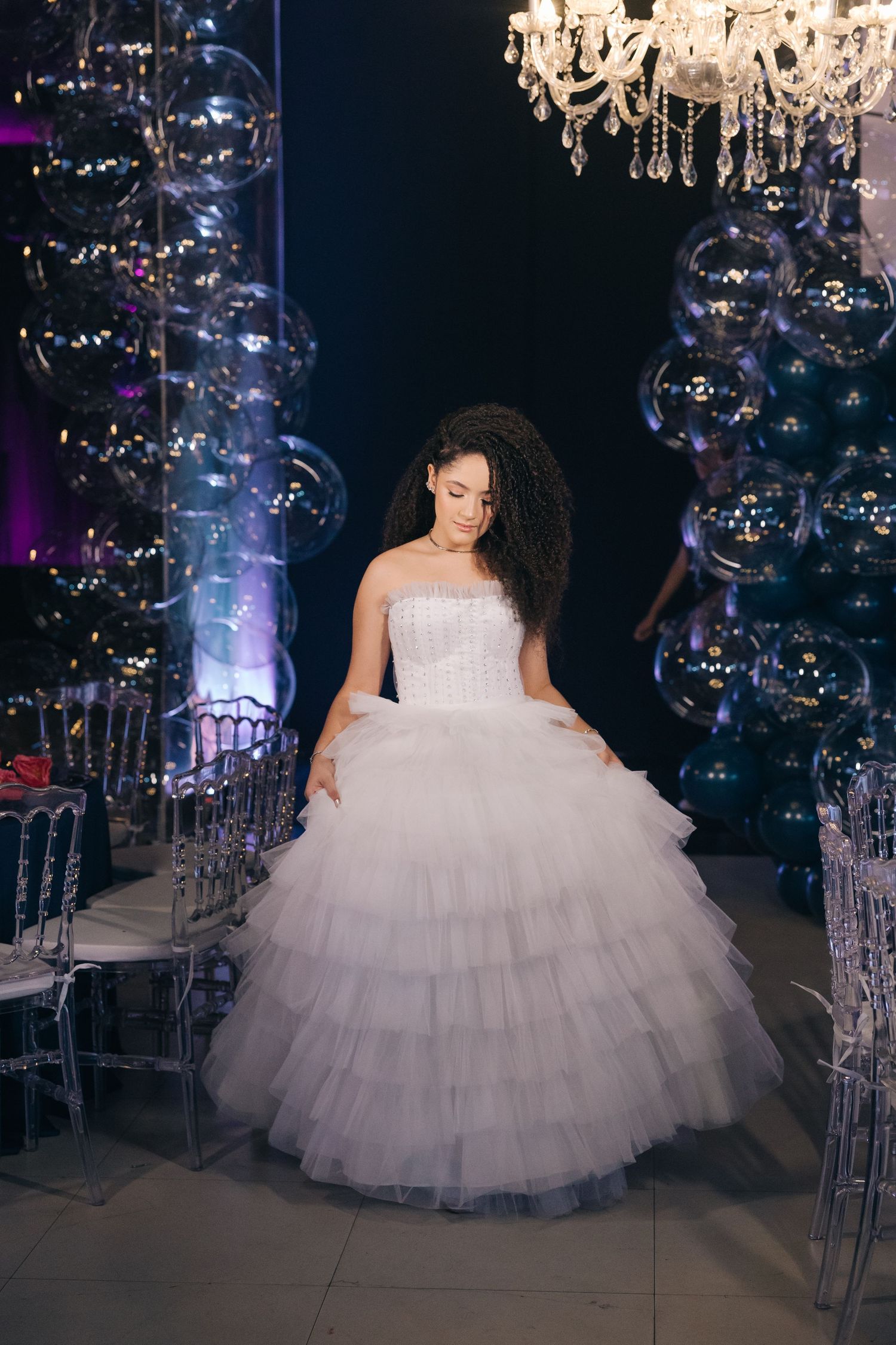 A layered tulle ballgown is showcased in an elegant setting with crystal chandeliers and bubbles creating a magical atmosphere.