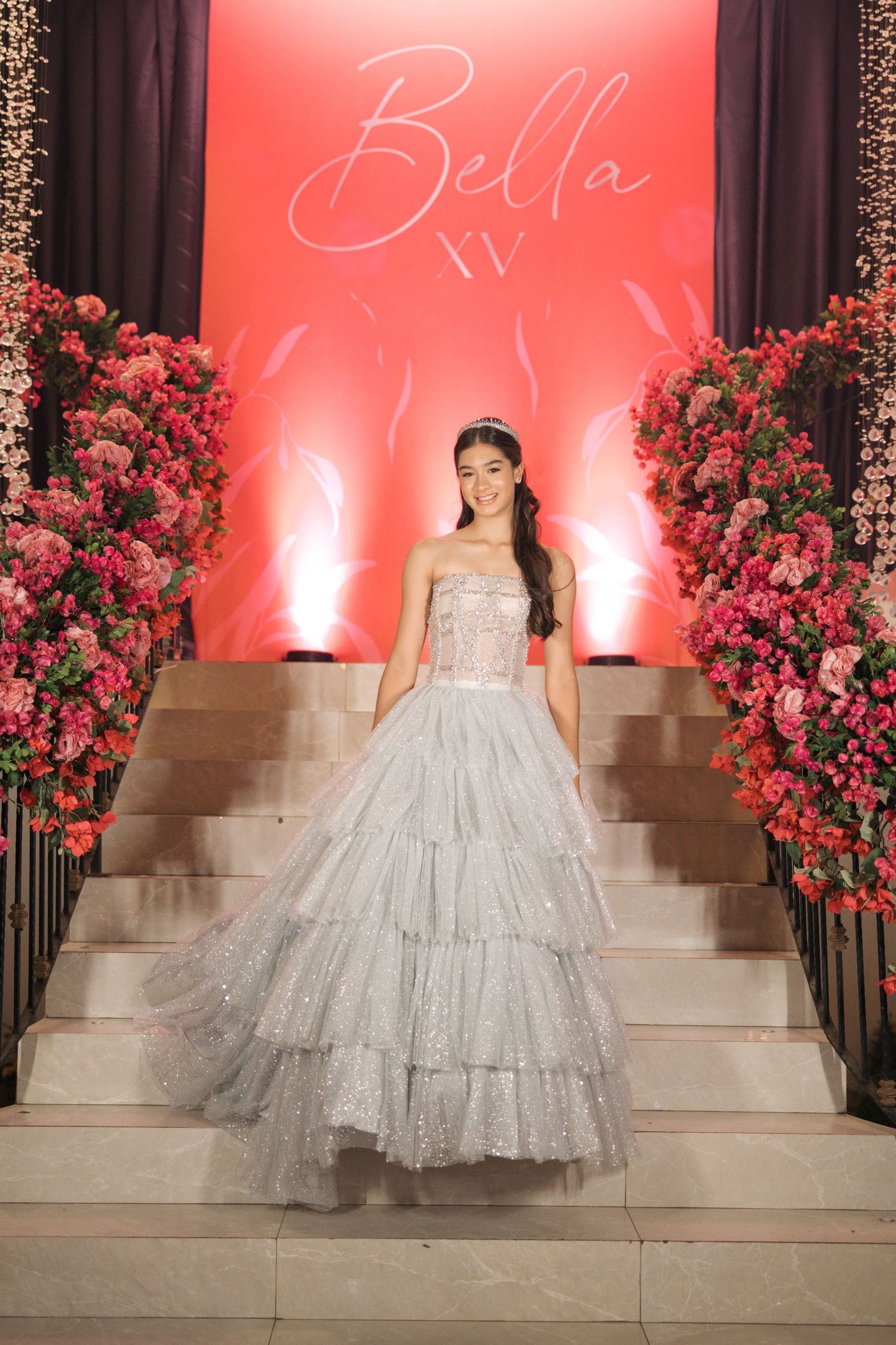 A stunning silver ballgown on display against a red backdrop with pink floral arrangements on grand staircase.
