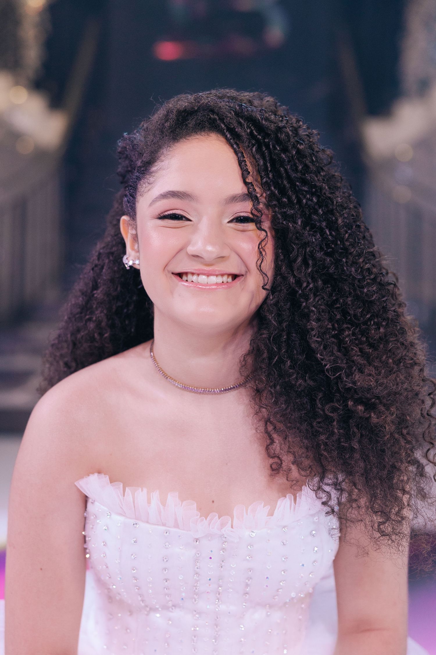 A close up portrait showing curly hair, jewelry and white formal dress details.