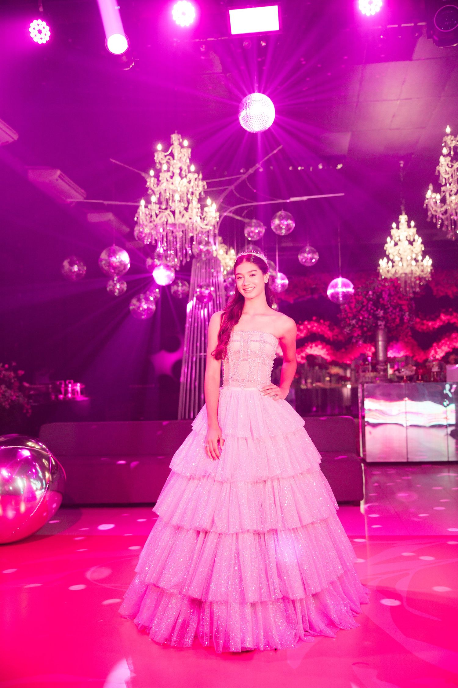 A tiered ballgown glows in pink lighting beneath crystal chandeliers at an elegant event.