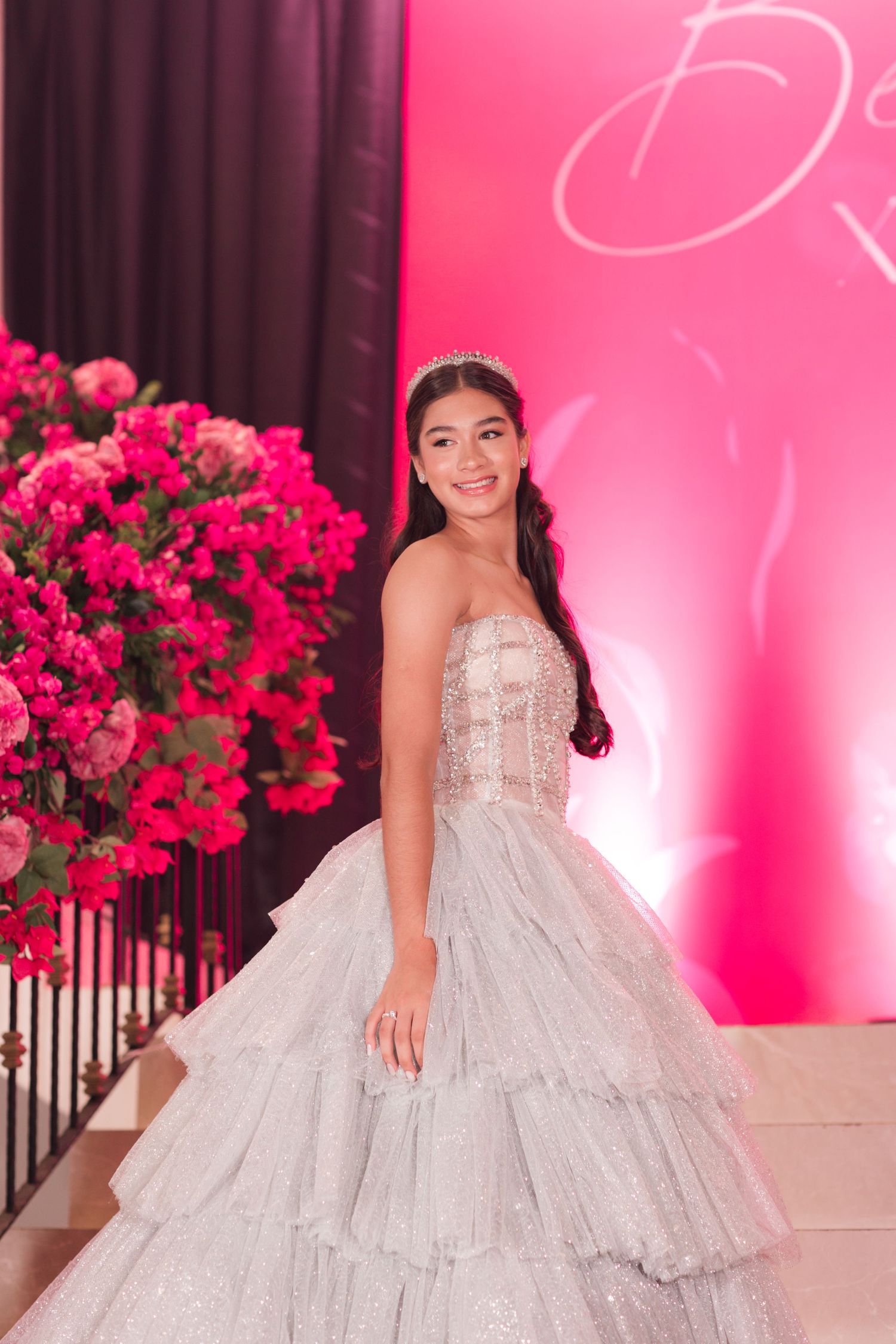 A young woman in a stunning tiered tulle ballgown poses in front of a pink backdrop with vibrant red floral arrangements.