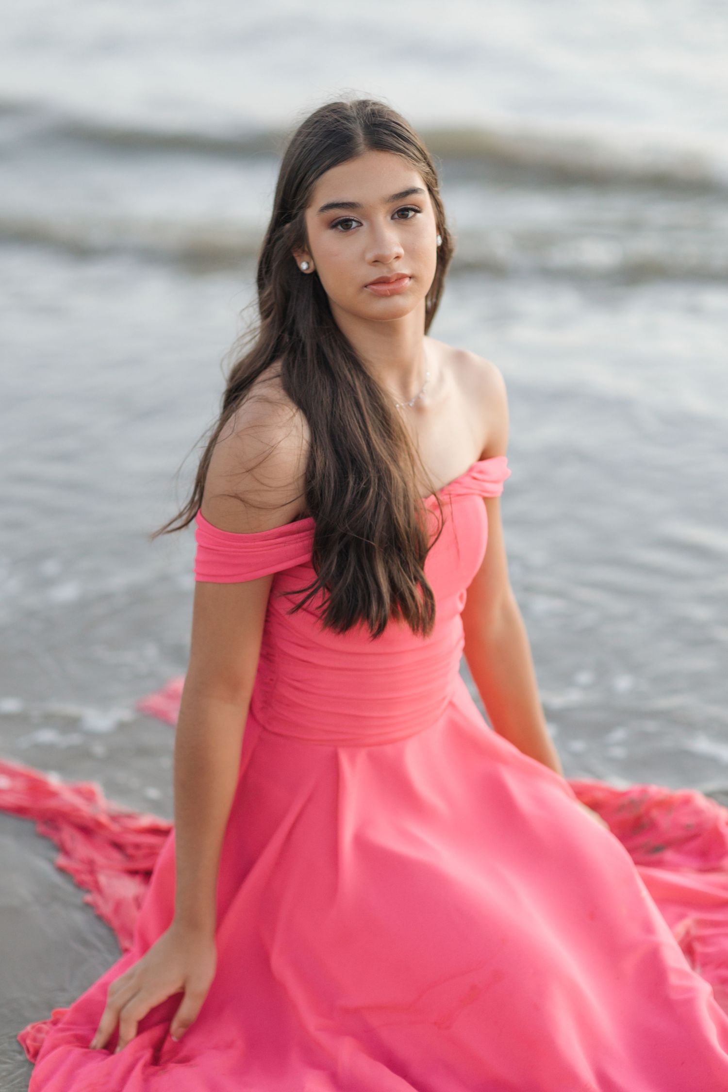 Elegant coral off-shoulder gown photographed along ocean shoreline at dusk.
