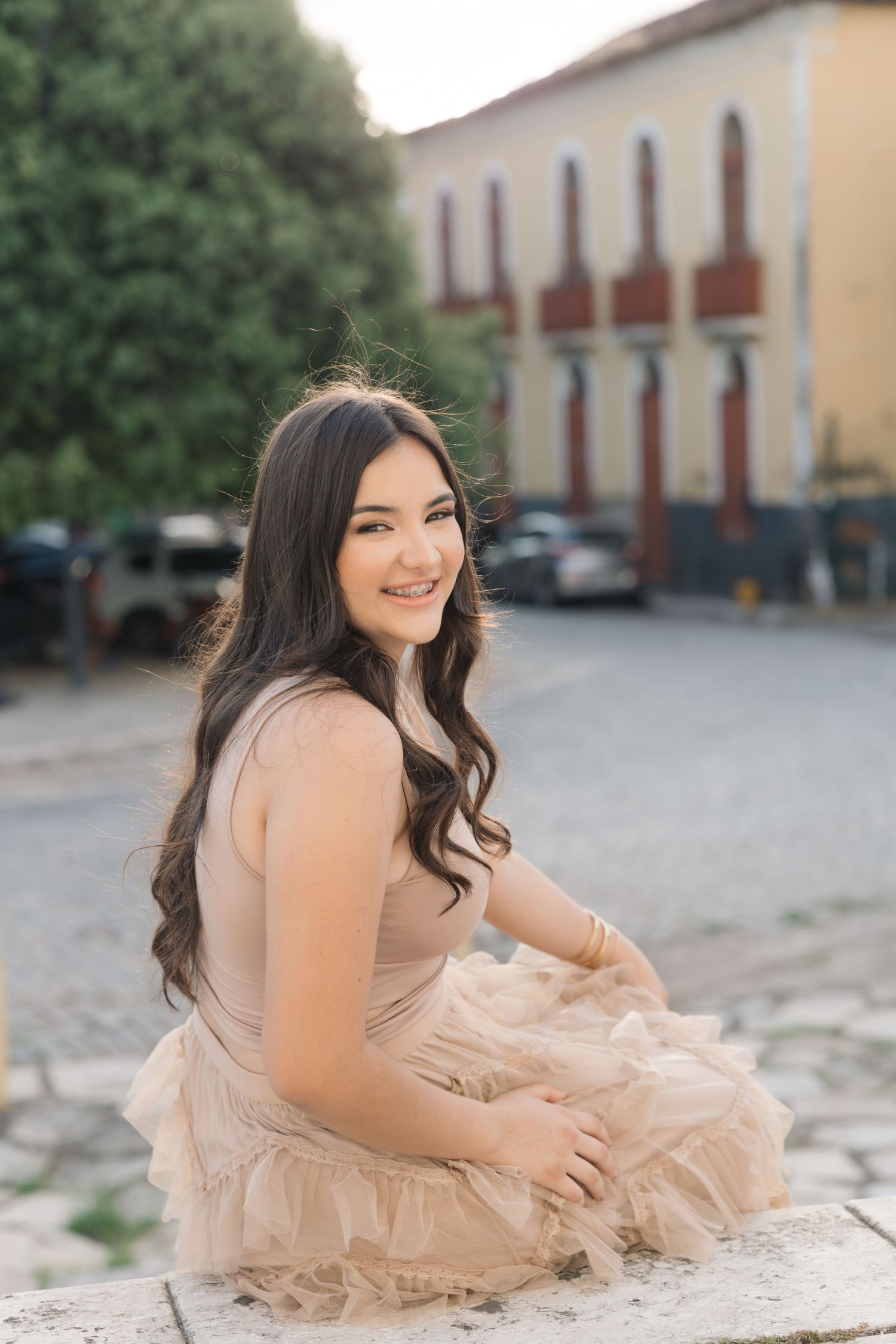 A person in a beige tulle dress sits on stone steps near a colonial style building.