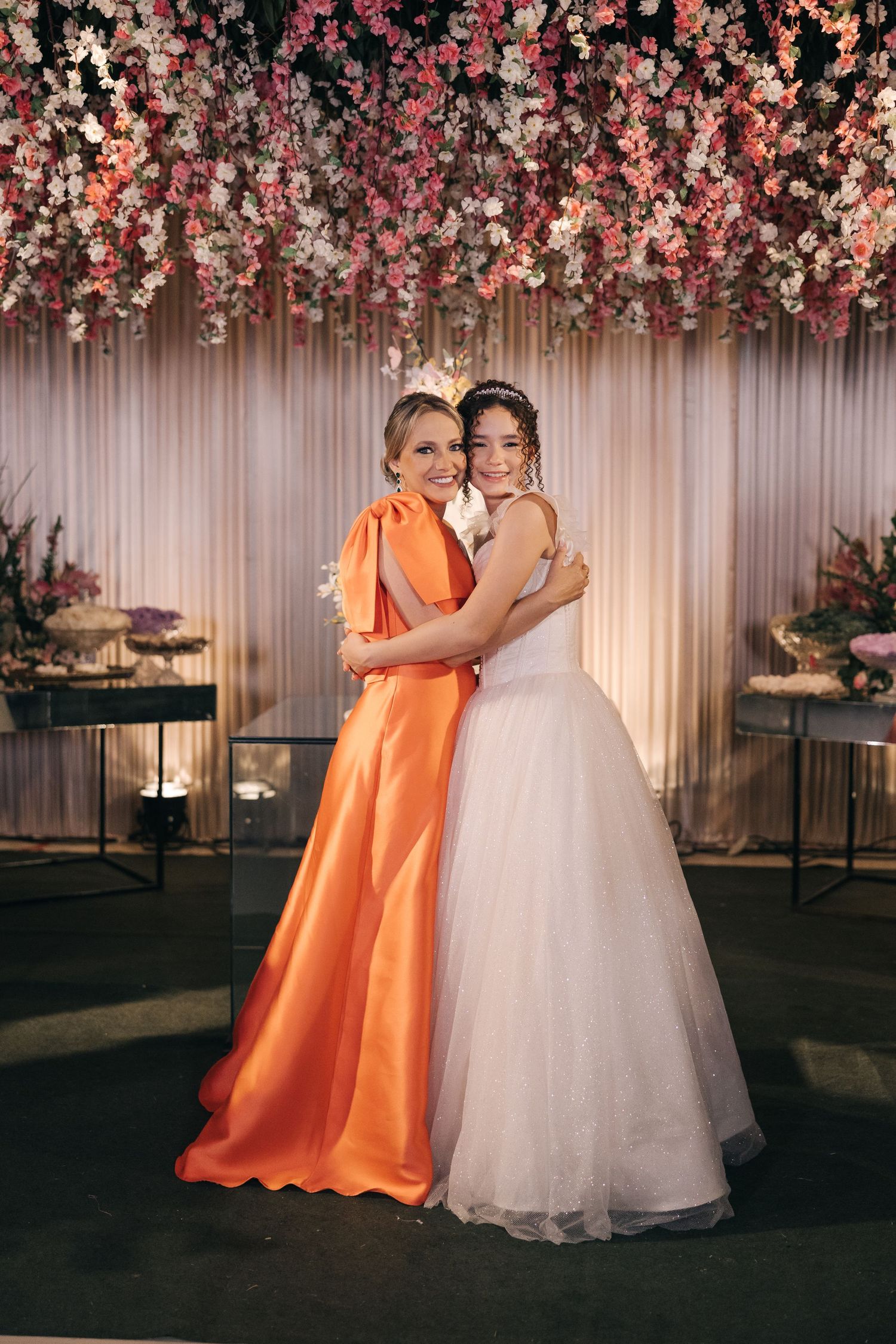 Two people pose together at a wedding celebration under a floral canopy, one in orange dress and one in white gown.