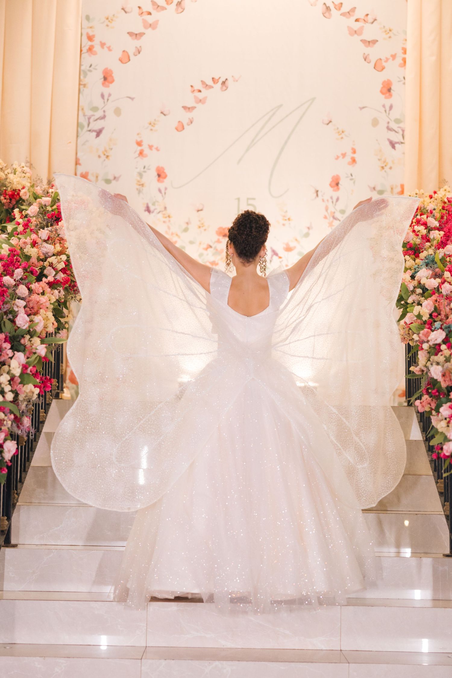A bride in a flowing white ballgown spreads her dress like wings against a backdrop of pink floral arrangements.