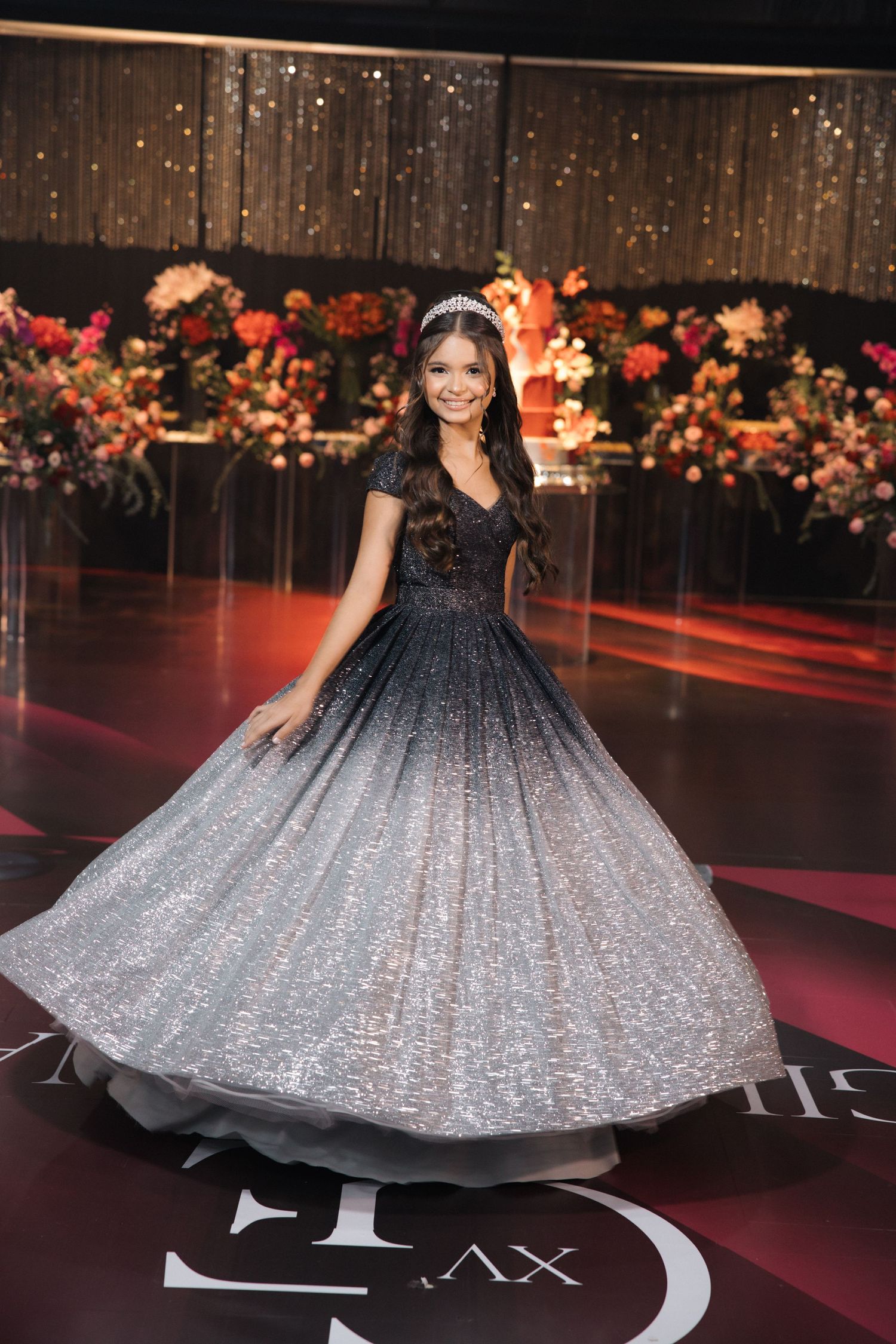An elegant ombre gown with sparkles twirls on a red-lit stage decorated with floral arrangements.
